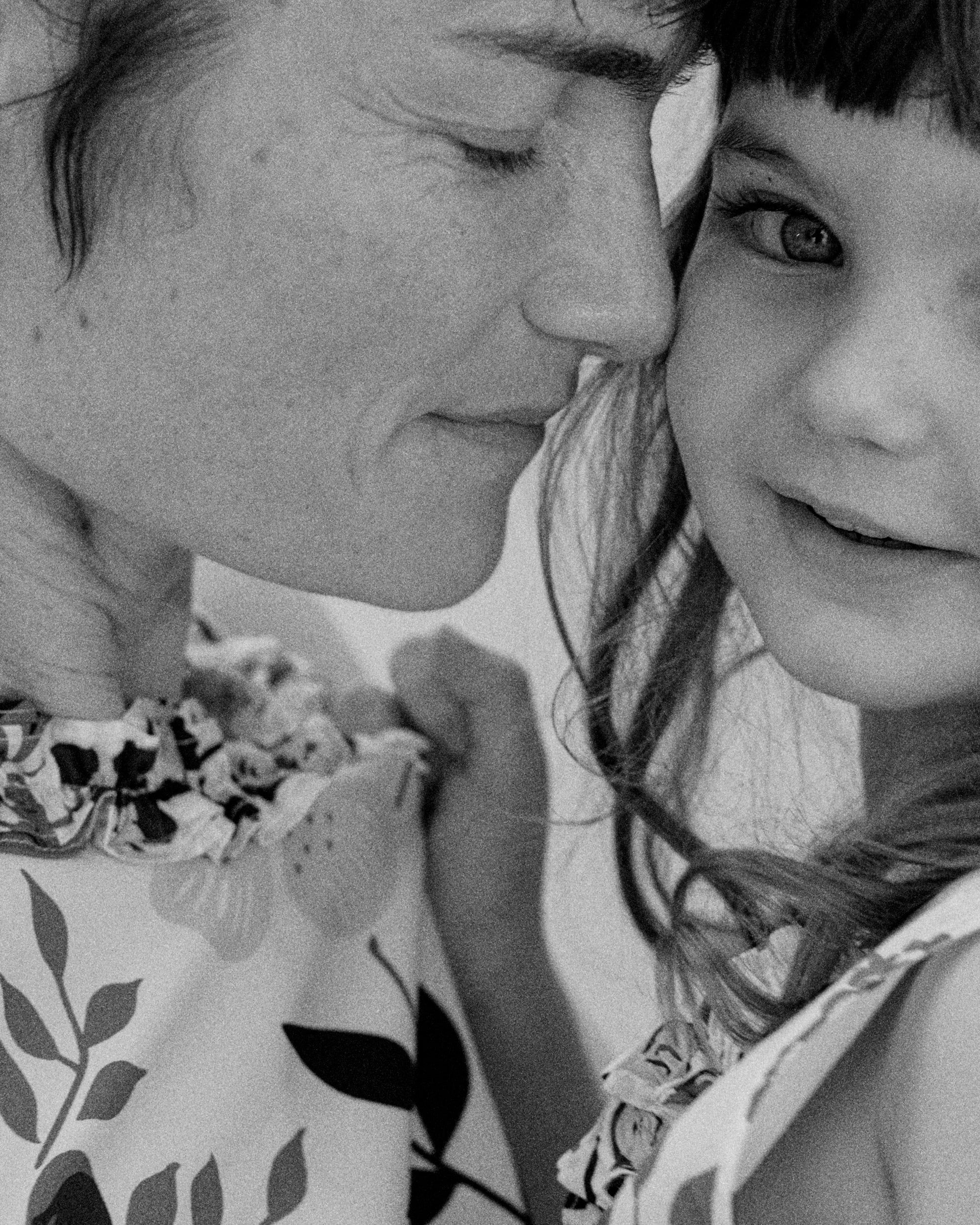 Black and white close-up portrait of two people — an adult and a child — cheek to cheek in a tender, intimate moment; both wearing light garments with delicate floral and botanical prints in the SS26 Liberty collection