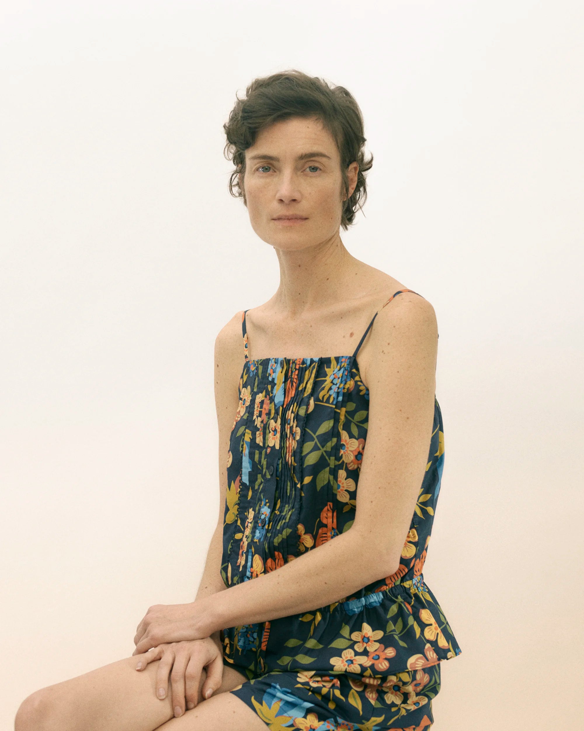 Female model seated and looking directly at the camera, wearing a spaghetti-strap camisole top in lustrous satin-like fabric with a gathered centre front and a rich navy ground printed with an exuberant Liberty floral motif featuring yellow, orange and green tropical blooms and foliage — paired with matching high-waisted shorts with an elasticated waist, creating a fluid, relaxed co-ord silhouette against a soft white background