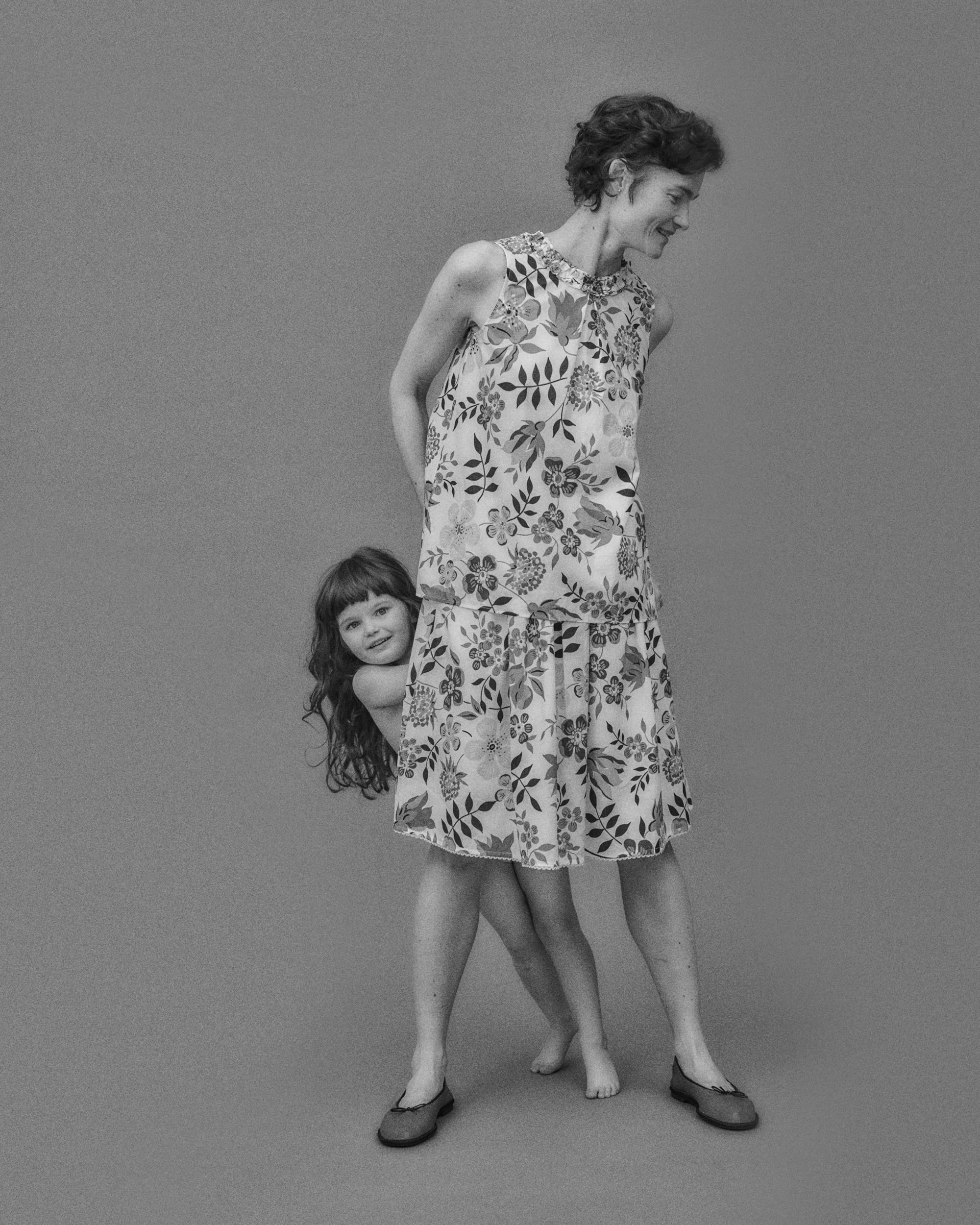 Black and white photograph of an adult woman and a young girl playfully posed together; the woman wears a sleeveless floral top with a ruffle neckline and a matching drop-waist skirt in lightweight fabric with an all-over Liberty botanical print, styled with flat ballet shoes — the child peeks out from behind her, barefoot and smiling, against a neutral grey background
