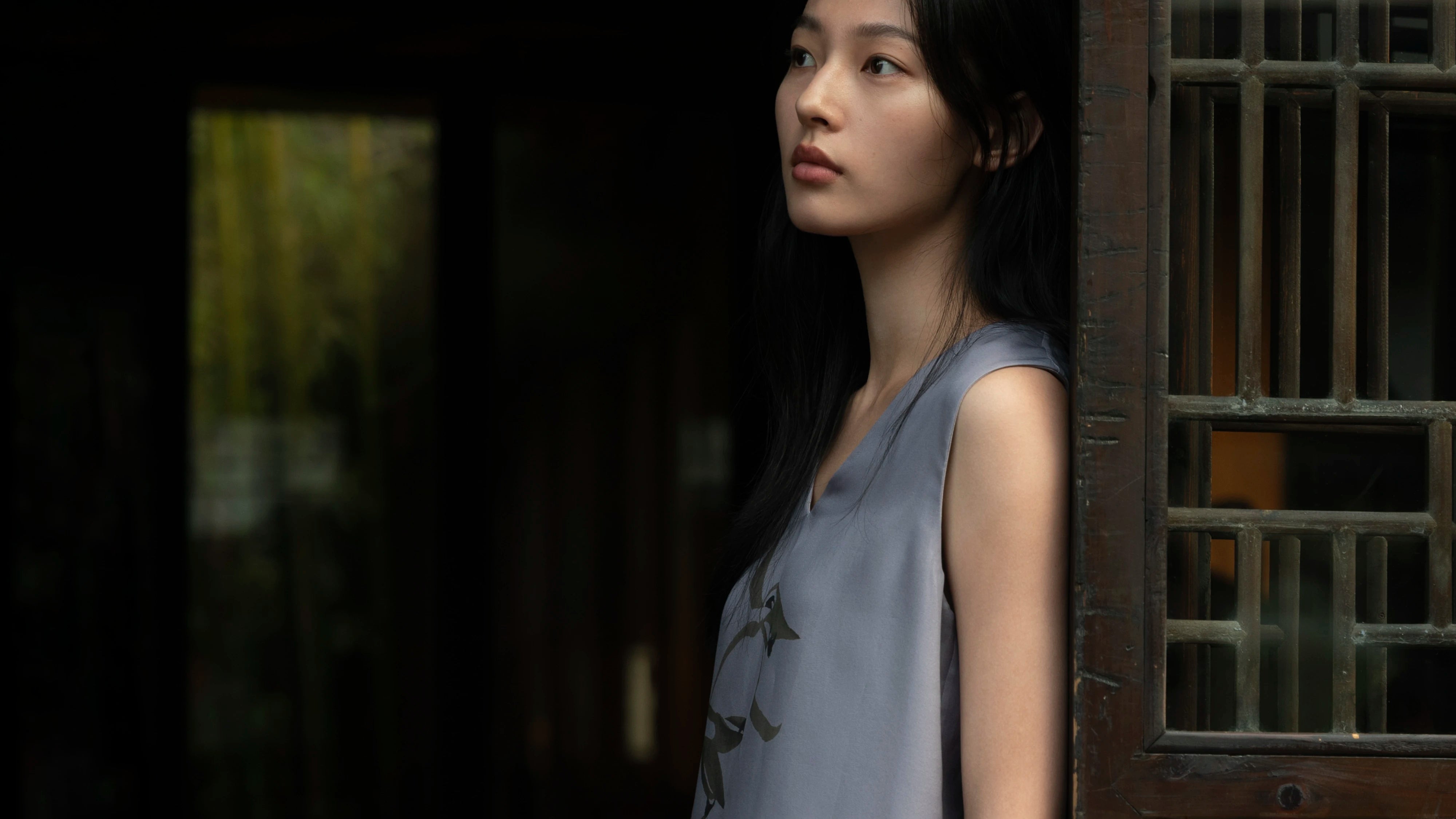 Female model photographed in profile in a dimly lit traditional interior, standing beside a dark wooden latticed door frame — wearing a sleeveless collarless shift dress in soft slate-blue fabric with a smooth, fluid drape and a subtle dark botanical print of trailing vine leaves along the front placket — a serene, contemplative silhouette in a moody, atmospheric setting