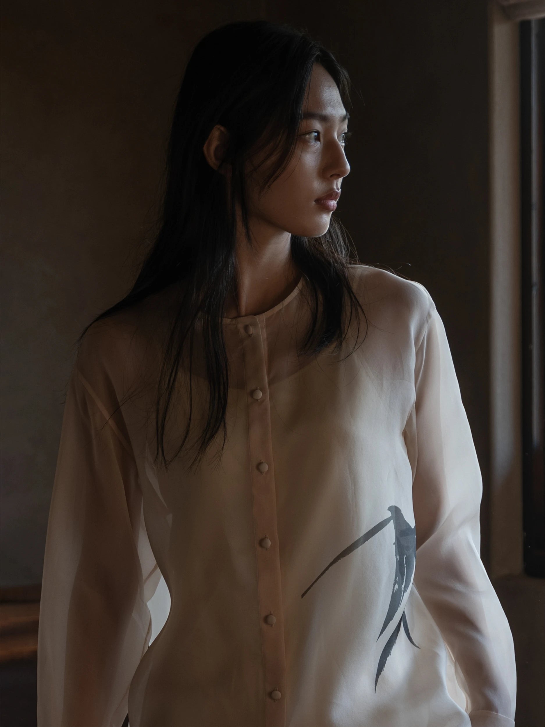 Female model photographed in three-quarter profile in a softly lit interior, wearing a long-sleeve mandarin-collar shirt dress in semi-sheer ivory fabric with a delicate, gauze-like texture and a fluid drape, fastened with small fabric-covered buttons down the centre front, featuring a single bold hand-painted dark ink brushstroke motif of a swallow in flight at the hip — a serene, minimalist silhouette with an artistic, calligraphic accent
