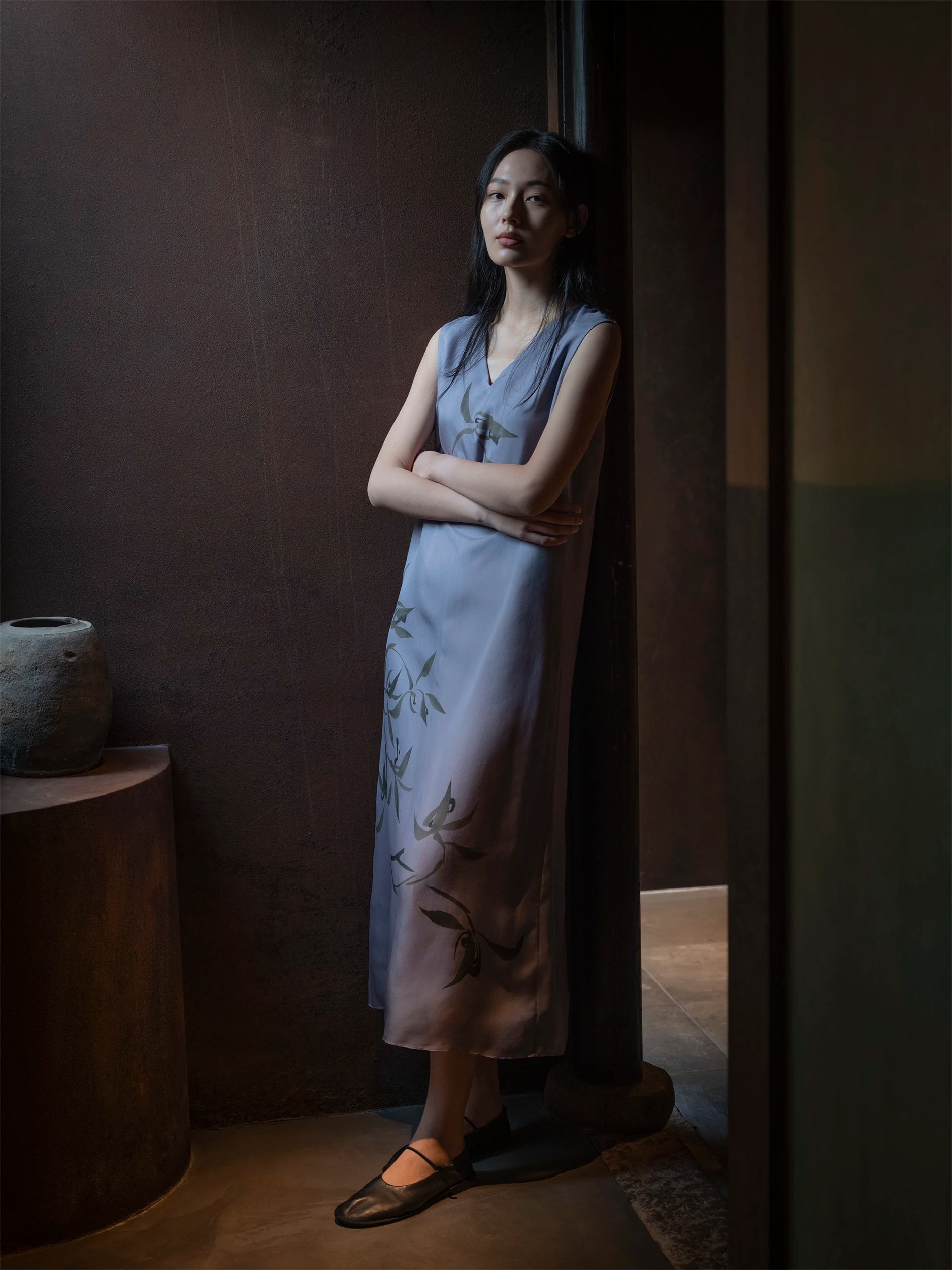 Female model standing with arms crossed, leaning against a dark wooden door frame in a moody, dimly lit interior — wearing a sleeveless V-neck midi dress in soft slate-blue fabric with a lustrous, silk-like drape and an all-over hand-painted botanical print featuring dark bamboo leaves and a small bird motif — a straight, elongated silhouette styled with dark brown leather Mary Jane flat shoes, beside a round wooden side table with a ceramic vase — a refined, contemplative mood