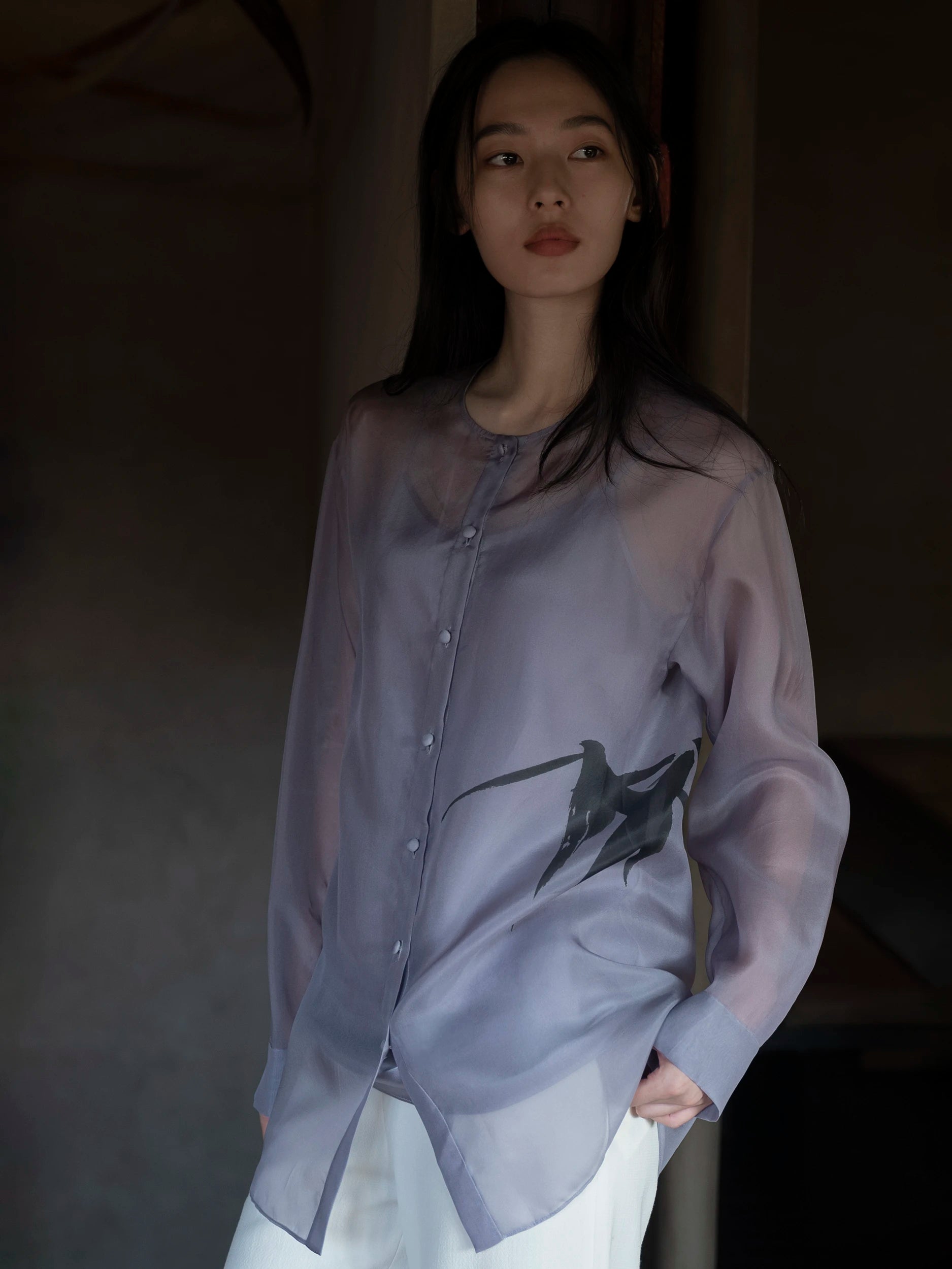 Female model standing in a dimly lit architectural interior, wearing an oversized long-sleeve mandarin-collar shirt in semi-sheer dusty-lavender fabric with a luminous, organza-like texture and a fluid drape, fastened with small metallic buttons down the centre front, featuring a bold hand-painted dark ink brushstroke motif of a swallow in flight at the hip — paired with wide-leg white trousers — a poetic, ethereal silhouette with a calligraphic artistic accent