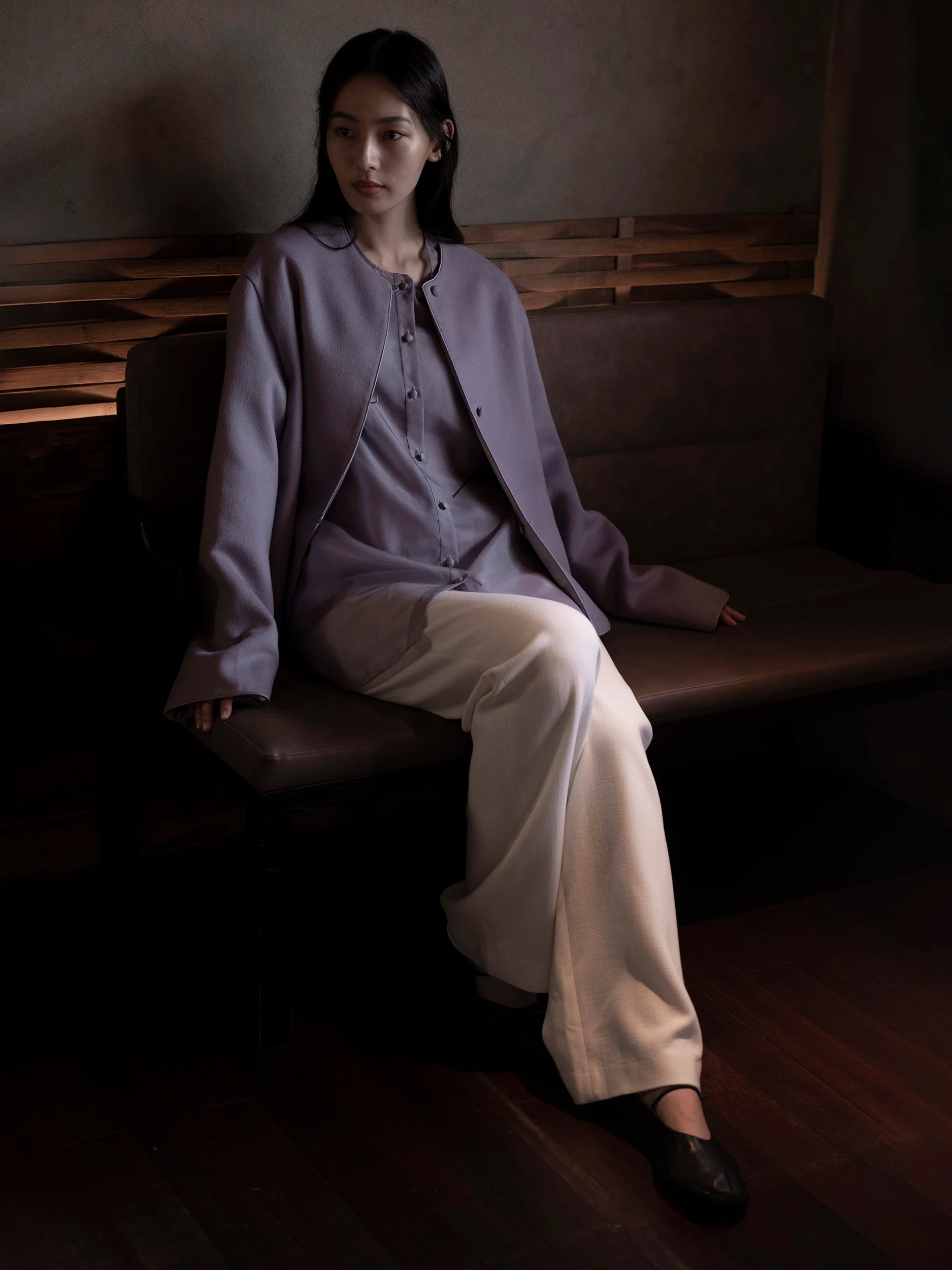 Female model seated on a dark wooden bench in a dimly lit interior, wearing an oversized collarless jacket in soft dusty-lavender woven fabric with a subtle texture, dark contrast piping along the edges and small dark buttons down the centre front, layered open over a matching lavender mandarin-collar shirt with a relaxed drape — paired with wide-leg ivory trousers in smooth, fluid fabric with a clean straight silhouette, and dark leather Mary Jane flat shoes — a serene, tonal monochromatic look