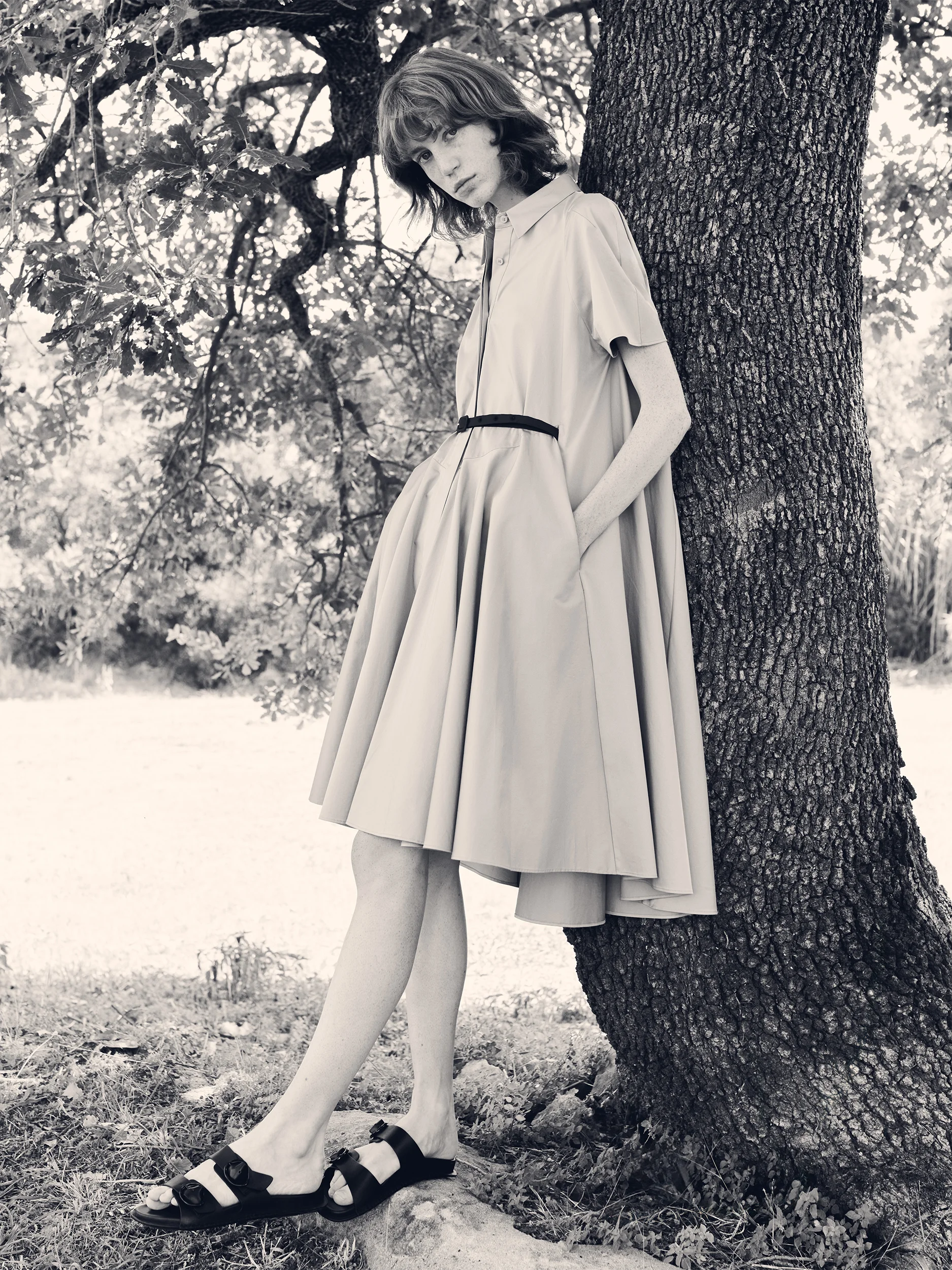 Black and white photograph of a female model leaning against a large tree trunk in a dappled Provençal landscape — wearing a voluminous short-sleeve shirt dress in crisp, structured cotton with a point collar, button-through front and a dramatically full pleated midi skirt, cinched at the waist with a thin dark grosgrain belt, hands in side pockets — styled with double-strap flat sandals in dark leather — a sculptural, architectural silhouette with a timeless, romantic mood