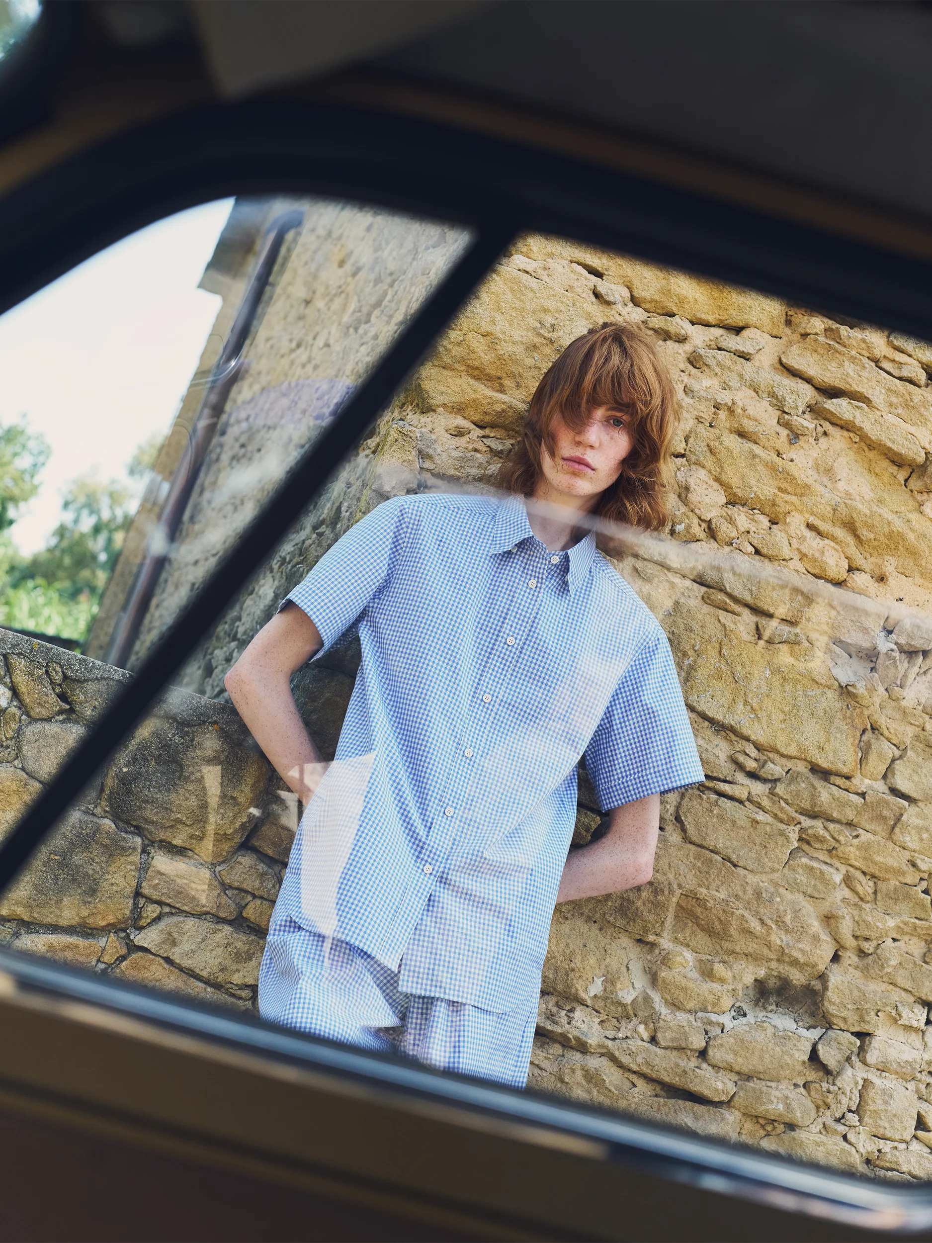 Female model photographed through a car window, leaning against a warm golden-stone wall in a Provençal village — wearing a matching co-ord set in blue-and-white gingham check cotton, consisting of an oversized short-sleeve shirt with a point collar and button front, paired with relaxed wide-leg shorts — a cool, effortless summer look with a vintage French countryside mood