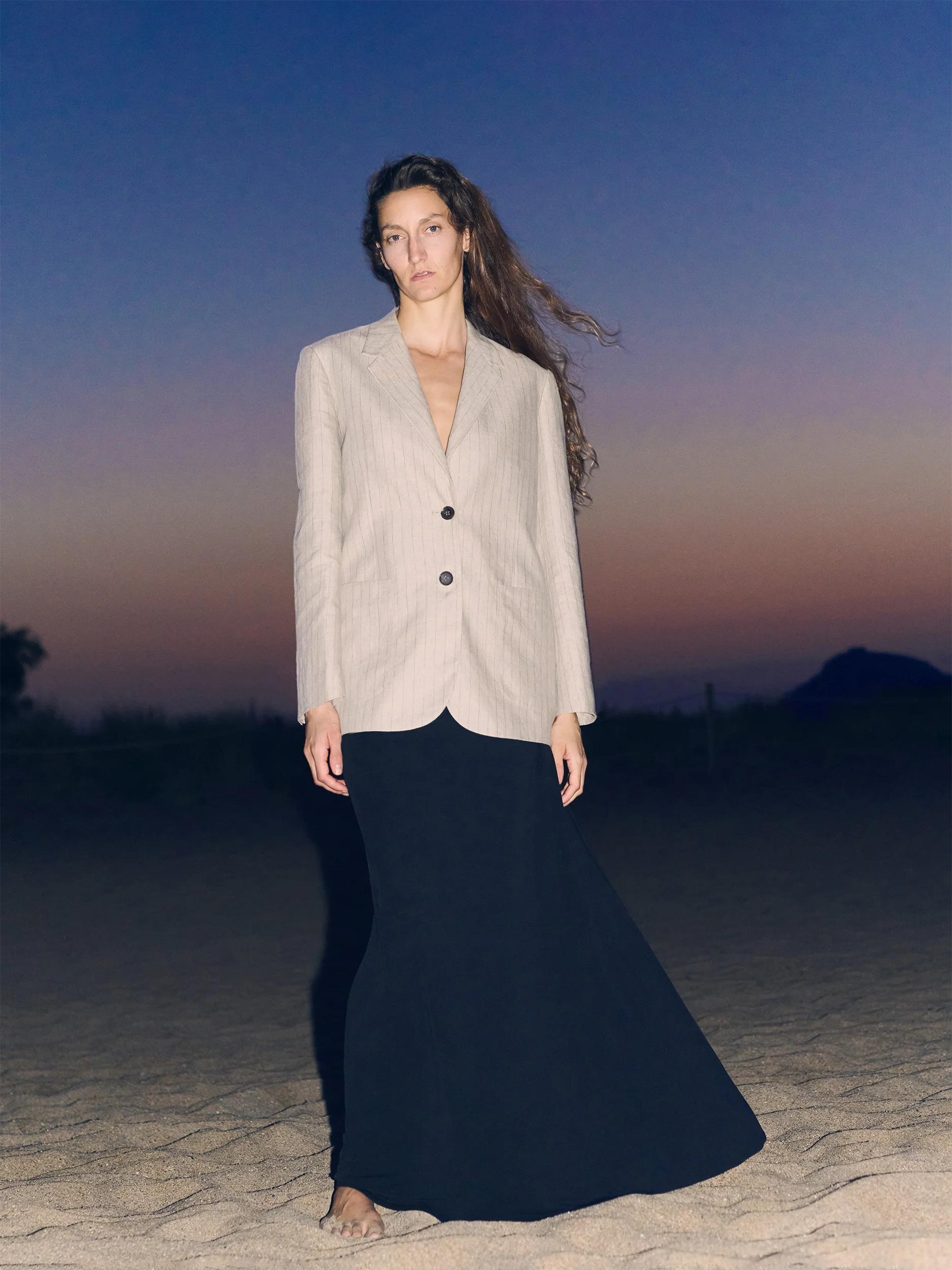 Female model standing barefoot on wind-swept sand dunes at dusk, hair streaming in the breeze — wearing a tailored single-breasted blazer in natural oatmeal linen with a subtle texture, notched lapels and two dark horn buttons, worn open over bare skin, paired with a fluid dark navy maxi skirt in lightweight linen with a gently flared, floor-grazing silhouette — a dramatic, cinematic look against a deep indigo and rose twilight sky with dark mountain silhouettes