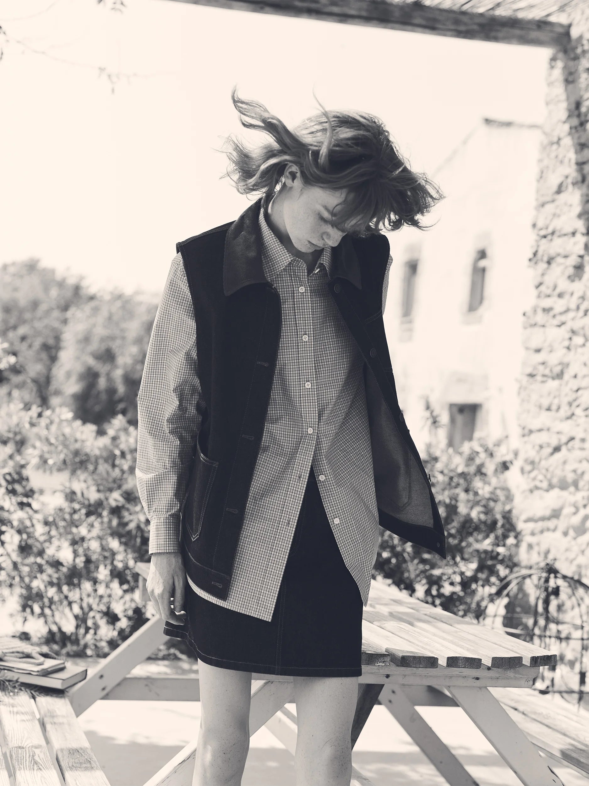 Black and white photograph of a female model with wind-swept hair, head bowed, standing beside a wooden picnic table on a sun-drenched stone terrace — wearing a longline sleeveless gilet in smooth dark fabric with leather-trimmed patch pockets and an open front, layered over an oversized long-sleeve shirt in fine gingham check cotton with a point collar and button front, paired with a dark mini skirt — a dynamic, effortlessly layered look with a Provençal countryside backdrop