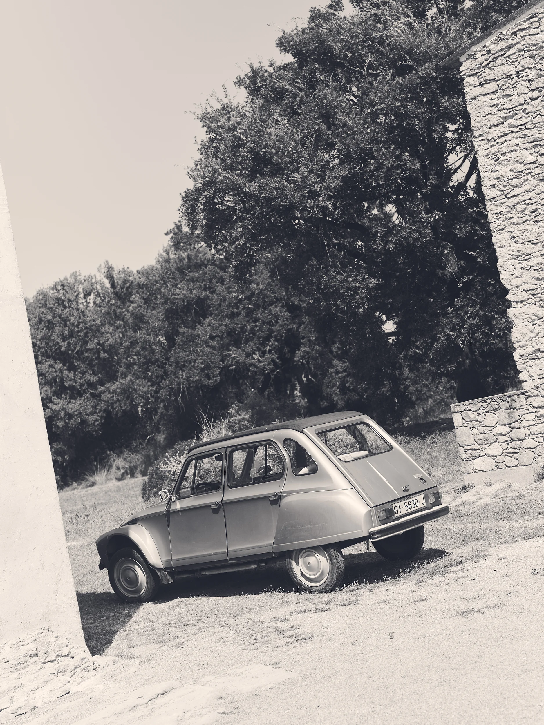 Black and white editorial photograph of a vintage Citroën Dyane parked on a sun-baked gravel courtyard beside a rustic dry-stone wall and tall leafy trees — a nostalgic, cinematic scene evoking the French countryside in summer, with no model or garments visible — used as an atmospheric mood image for the SS26 C-Line collection
