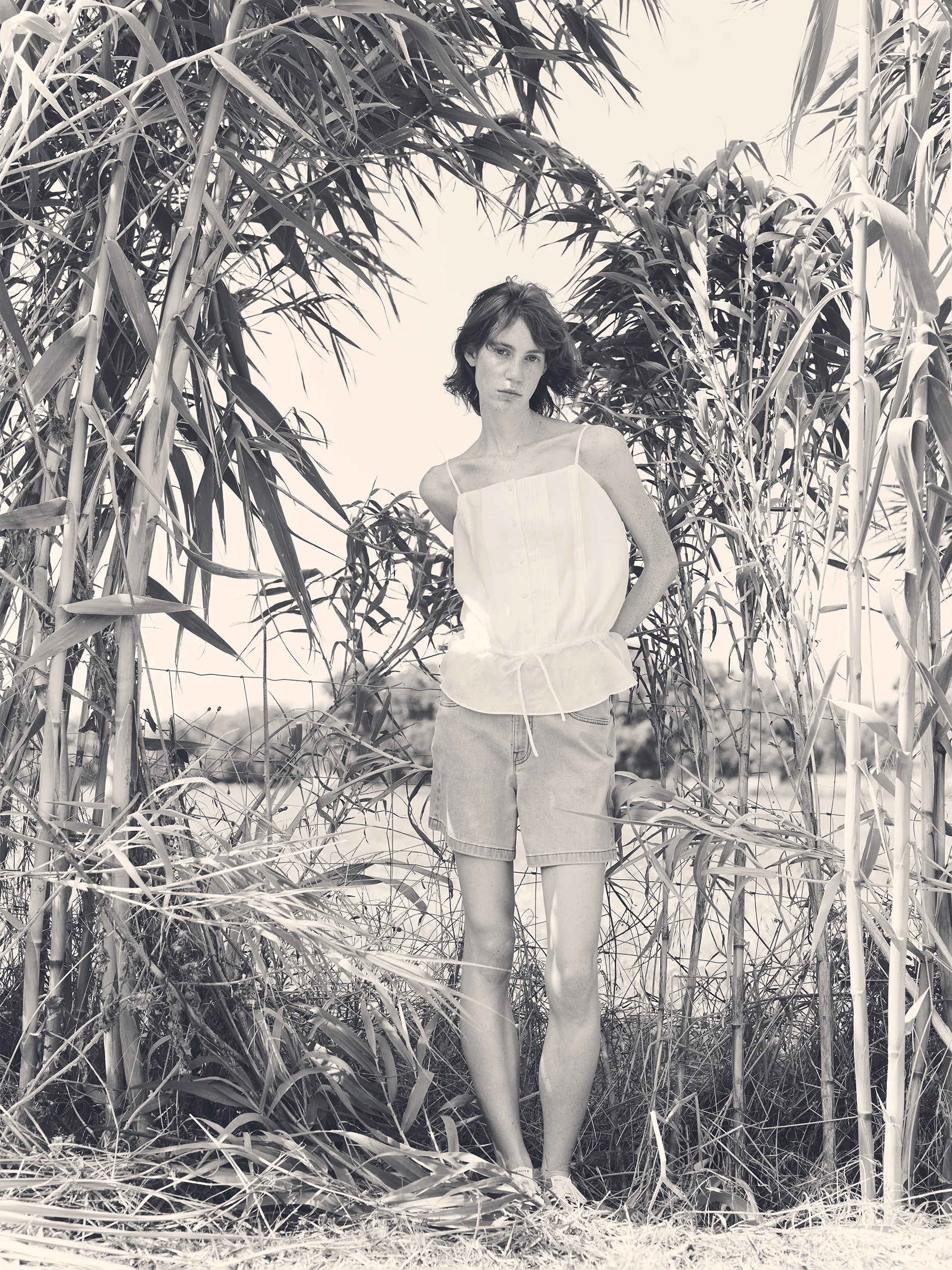 Black and white photograph of a female model standing confidently among tall bamboo and reed stalks in a wild outdoor setting — wearing a delicate spaghetti-strap camisole top in lightweight ivory fabric with a relaxed peplum silhouette and a thin drawstring tie at the waist, paired with straight-cut bermuda shorts in washed cotton with a clean, casual silhouette and flat canvas shoes — a serene, naturalistic editorial mood