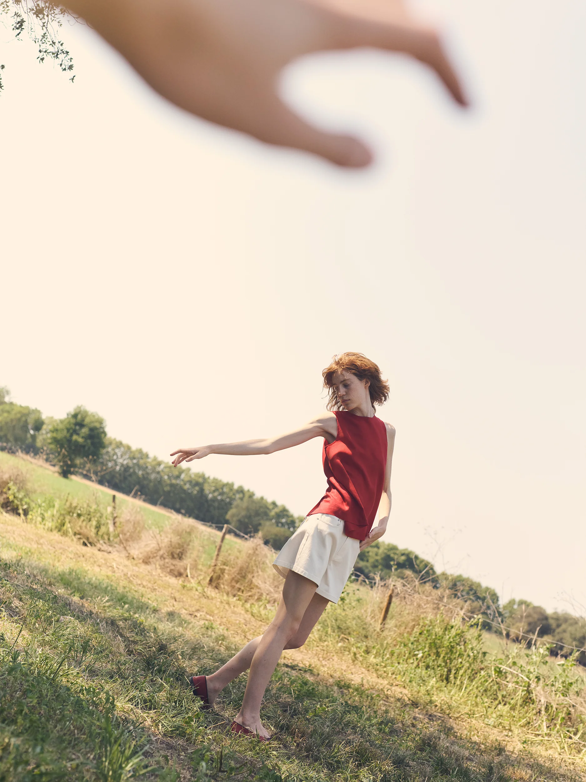 Female model dancing freely on a sun-drenched grassy hillside, arms outstretched — wearing a sleeveless crew-neck top in smooth crimson-red fabric with a clean, fitted silhouette, tucked into a flared mini skirt in crisp ivory fabric with a subtle A-line volume and small stud embellishments at the waistband, styled with dark burgundy flat Mary Jane shoes — a joyful, cinematic outdoor moment with a large hand reaching into frame from above against a hazy pale sky