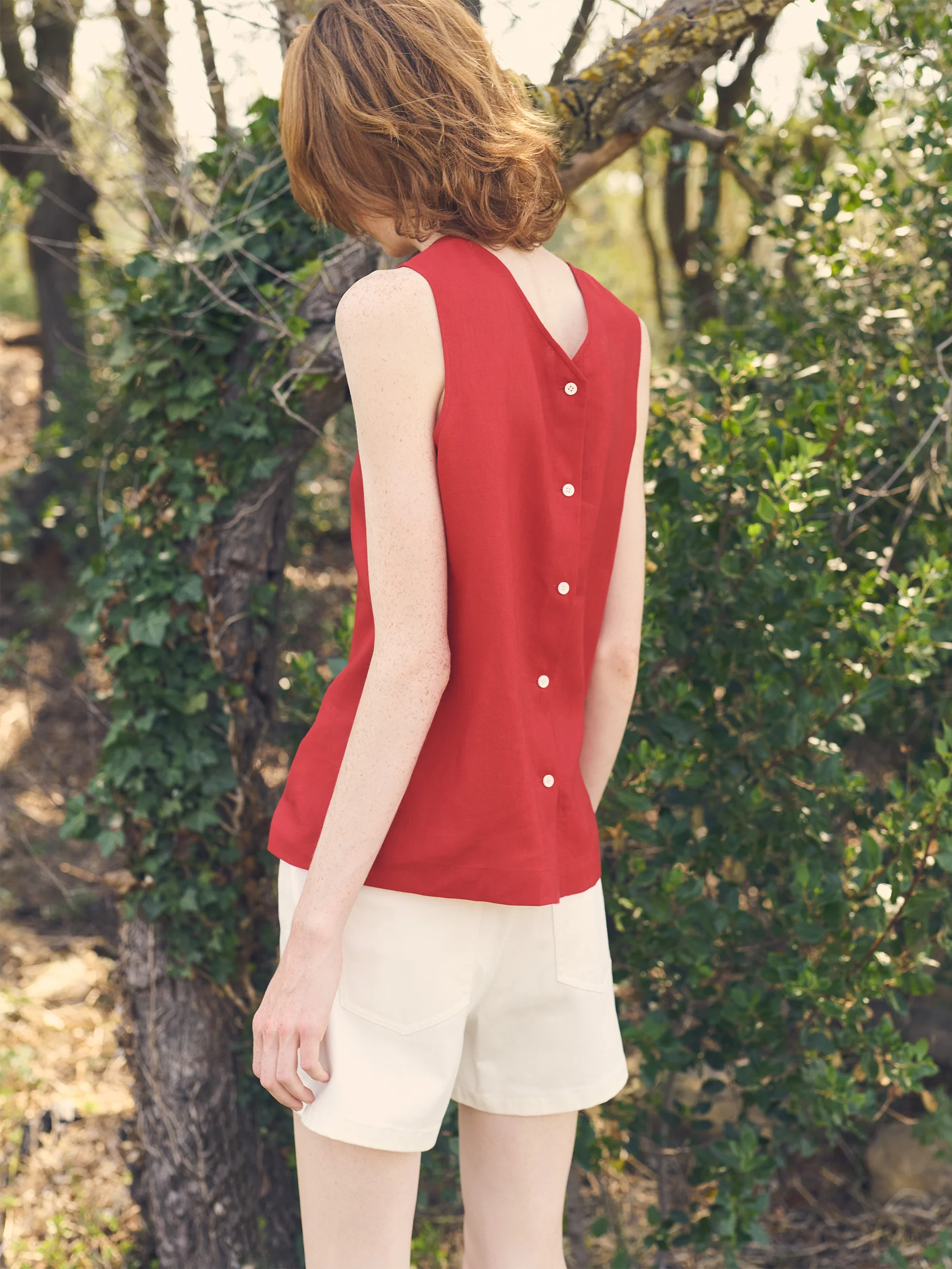 Female model photographed from behind in a sun-dappled Mediterranean garrigue landscape — wearing a sleeveless crew-neck top in smooth crimson-red fabric with a fitted, clean silhouette and a distinctive back button fastening with a row of small round pearlescent buttons running from neckline to hem, paired with tailored ivory mini shorts in crisp structured fabric — an elegant, refined detail shot highlighting the signature back design