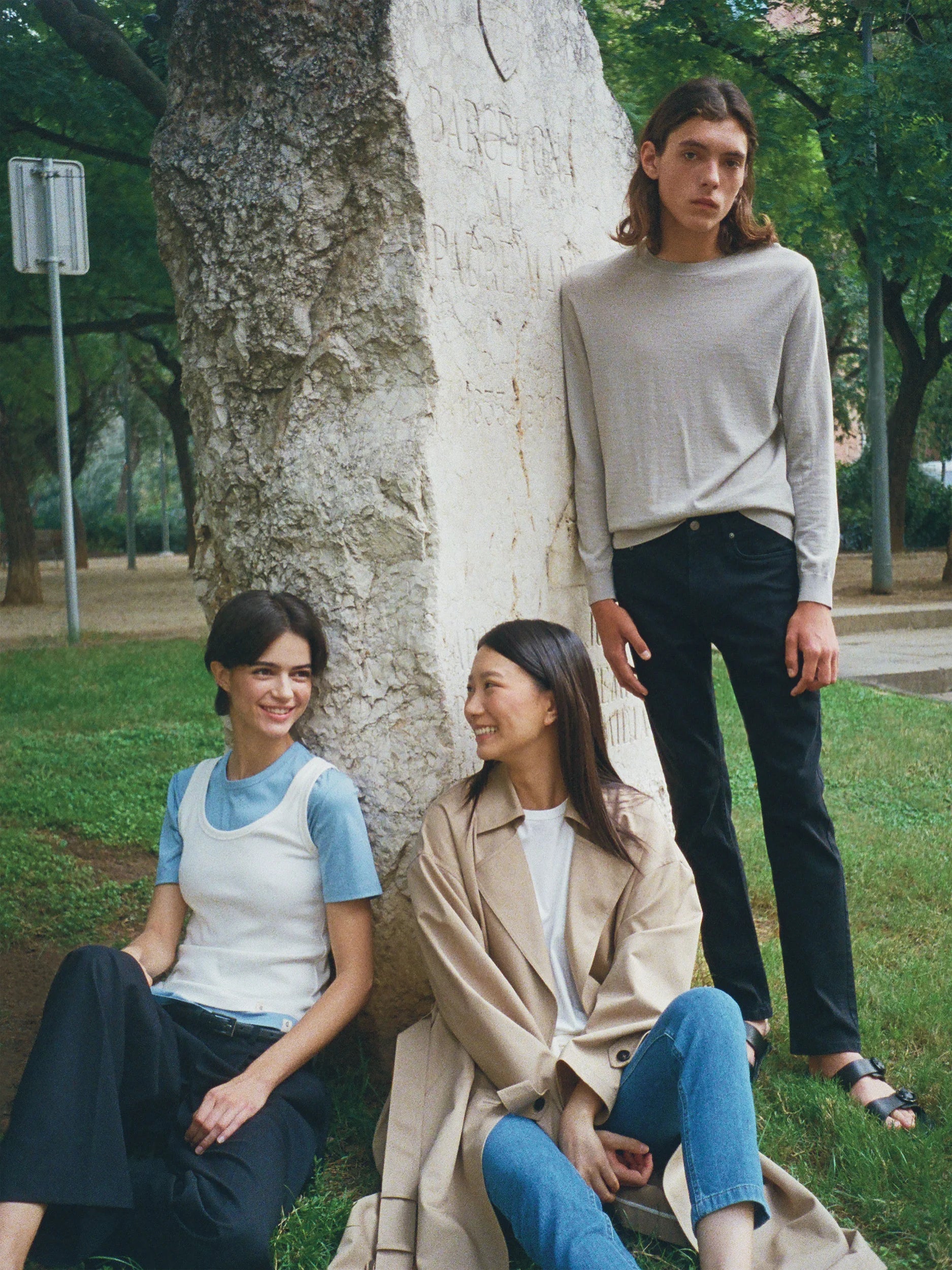 Three models gathered around a large pale-barked tree in a leafy urban park — a woman seated on the grass wearing a fitted white sleeveless tank top in smooth fine-knit jersey layered over a pale blue short-sleeve tee, paired with wide-leg black trousers; a woman seated leaning against the tree in a longline camel-beige trench coat in structured woven fabric with a double-breasted front and belt, worn open over a white tee and slim blue jeans; a man standing behind in a sage-grey long-sleeve crew-neck top