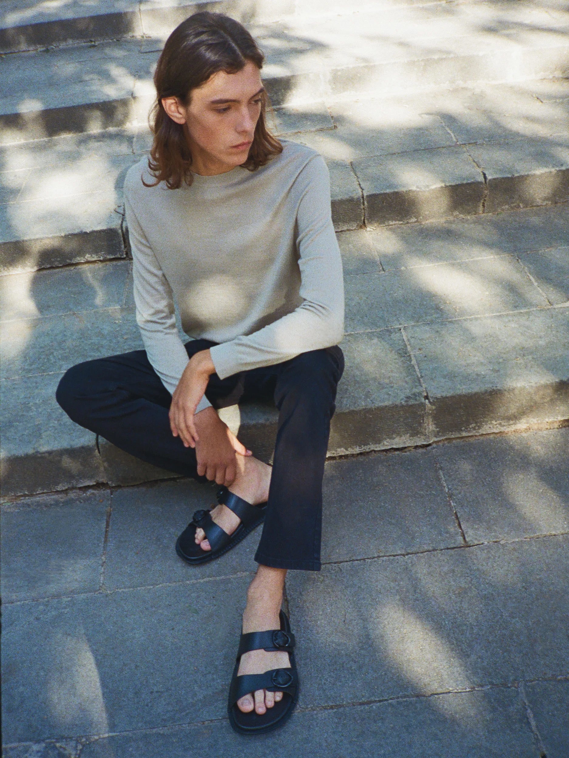 Male model seated on sun-dappled stone steps, wearing a fitted sage-grey long-sleeve crew-neck top in fine, semi-sheer lightweight jersey with a clean, body-skimming silhouette, paired with straight-cut cropped navy trousers and black leather double-strap flat sandals — a minimal, understated urban look in dappled natural light"