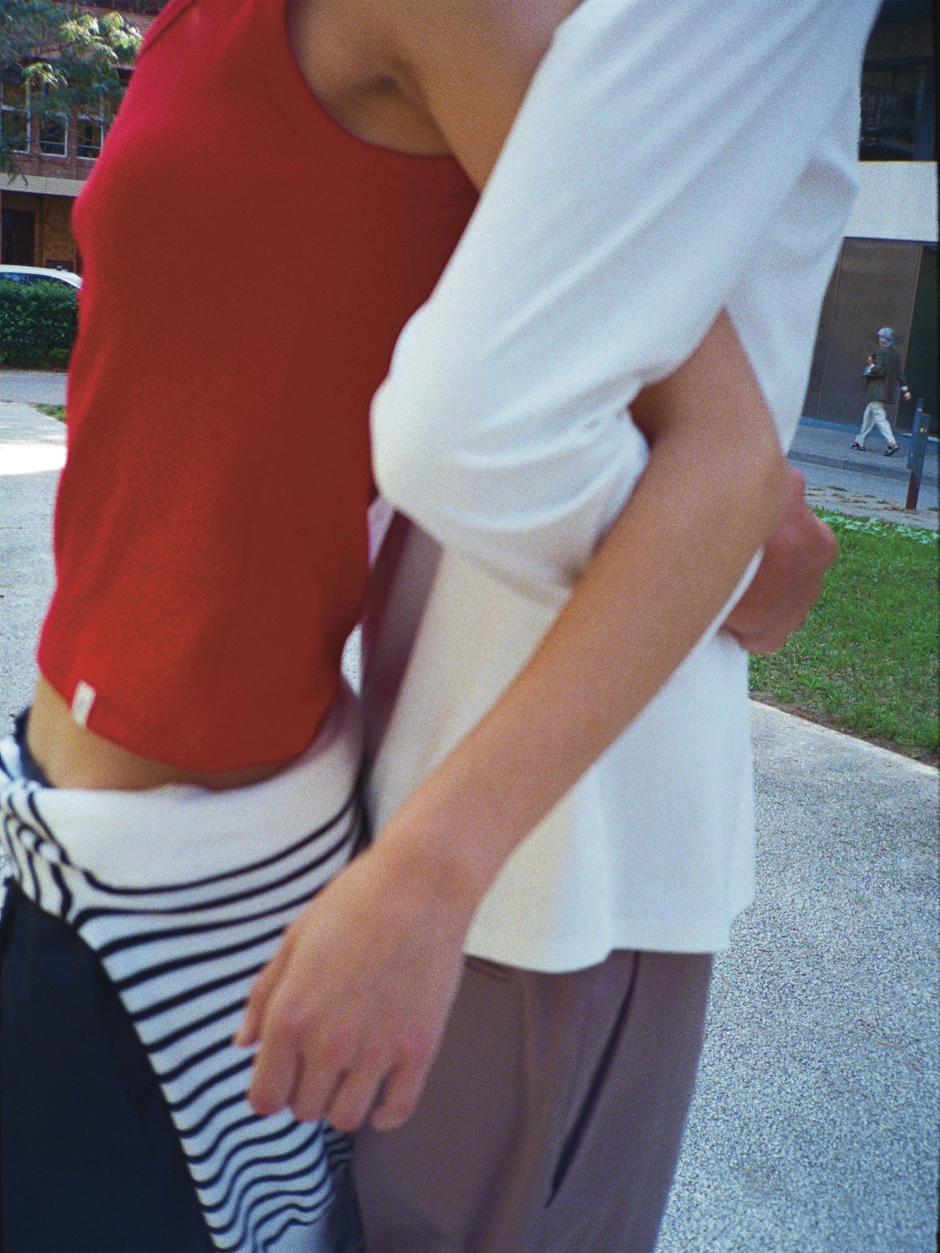 Close-up detail shot of two models embracing outdoors — one wearing a cropped scarlet red short-sleeve top in smooth fine-knit jersey with a relaxed fit, tucked into navy-and-white horizontal striped wide-leg trousers with a high waist; the other in a white short-sleeve top in soft cotton with a slightly flared hem, paired with taupe wide-leg trousers with a clean straight silhouette — an intimate, candid urban moment