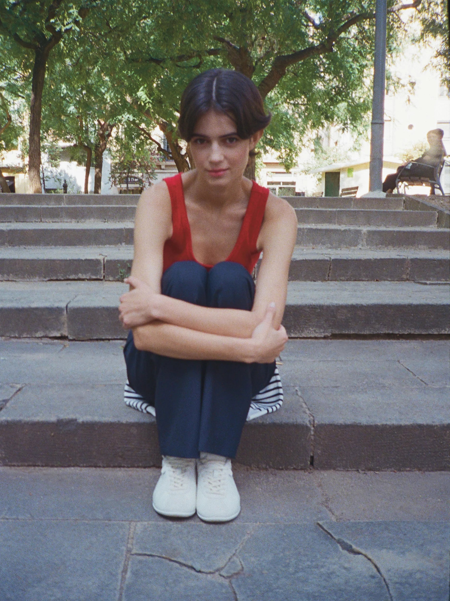 Female model seated on stone steps in an urban square, hugging her knees and looking directly at the camera — wearing a scarlet red sleeveless tank top in smooth fine-ribbed jersey with a deep scoop neckline and a fitted silhouette, layered over a navy-and-white horizontal striped top, paired with straight-cut navy trousers and white canvas lace-up sneakers — lush green trees and a sunlit city square in the background