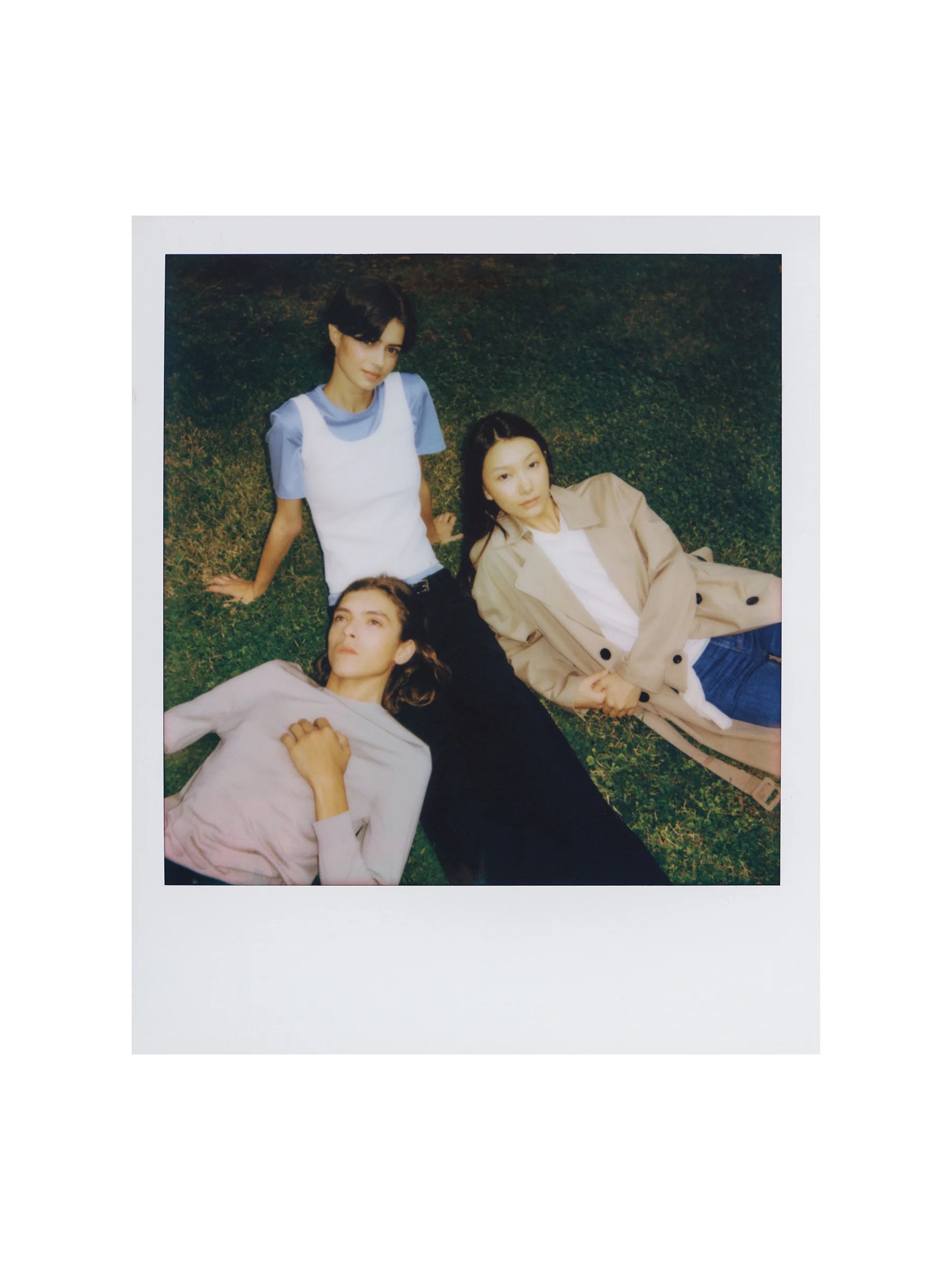 Polaroid-style photograph of three female models lying and lounging on green grass, shot from above — one in a fitted white short-sleeve crew-neck tee in smooth cotton jersey layered over a pale blue long-sleeve top; one in a soft mauve-pink long-sleeve sweatshirt in fine brushed jersey with a relaxed silhouette; one in a camel-beige oversized blazer in structured woven fabric with notched lapels and button front, worn over a white tee and blue denim shorts — a candid, nostalgic editorial mood