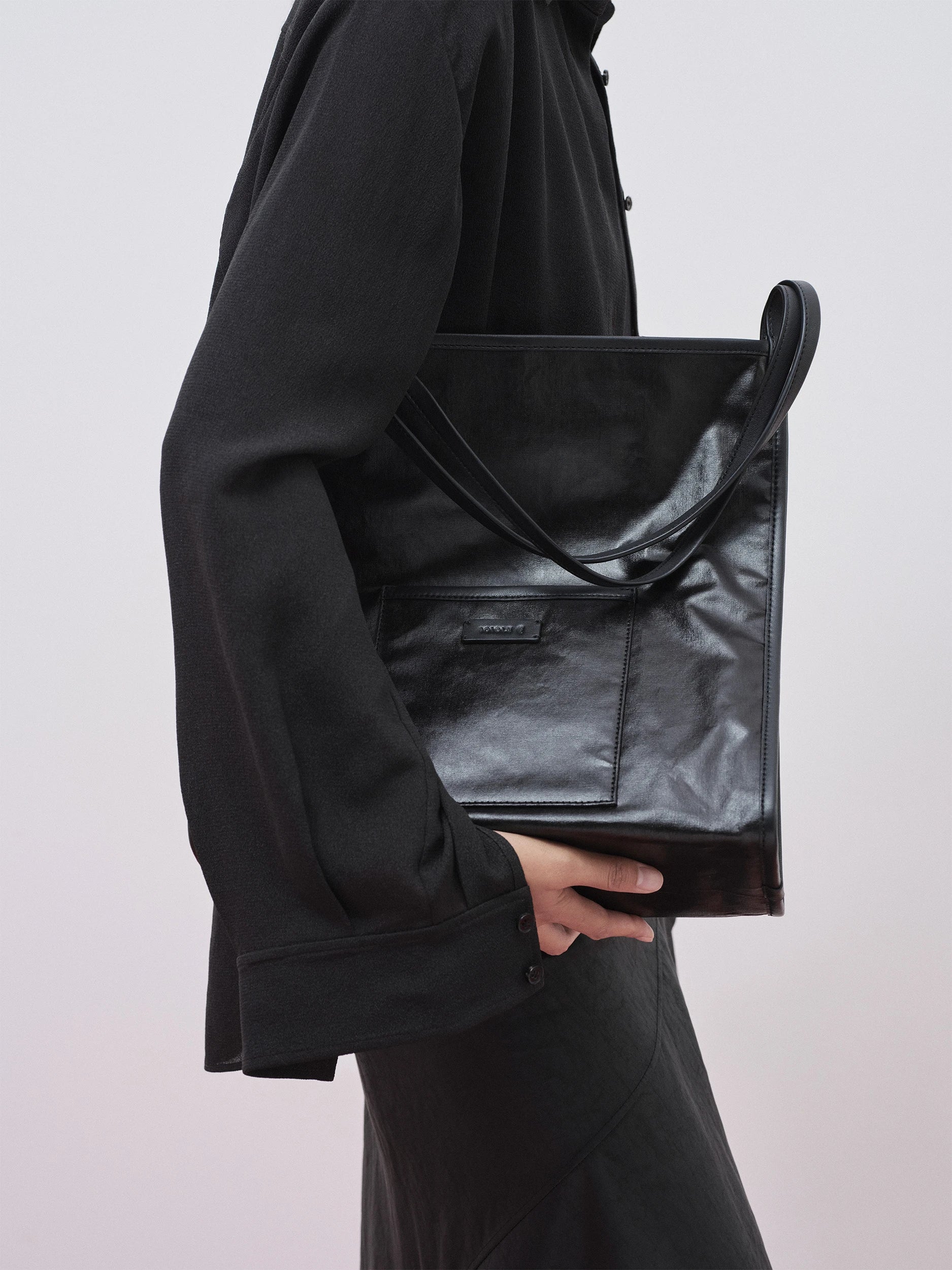 Close-up of a model holding a structured black leather tote bag with a slightly crinkled, supple texture, flat shoulder straps and a front patch pocket with a discreet brand label — carried against a black fluid gauze coat, against a neutral grey background