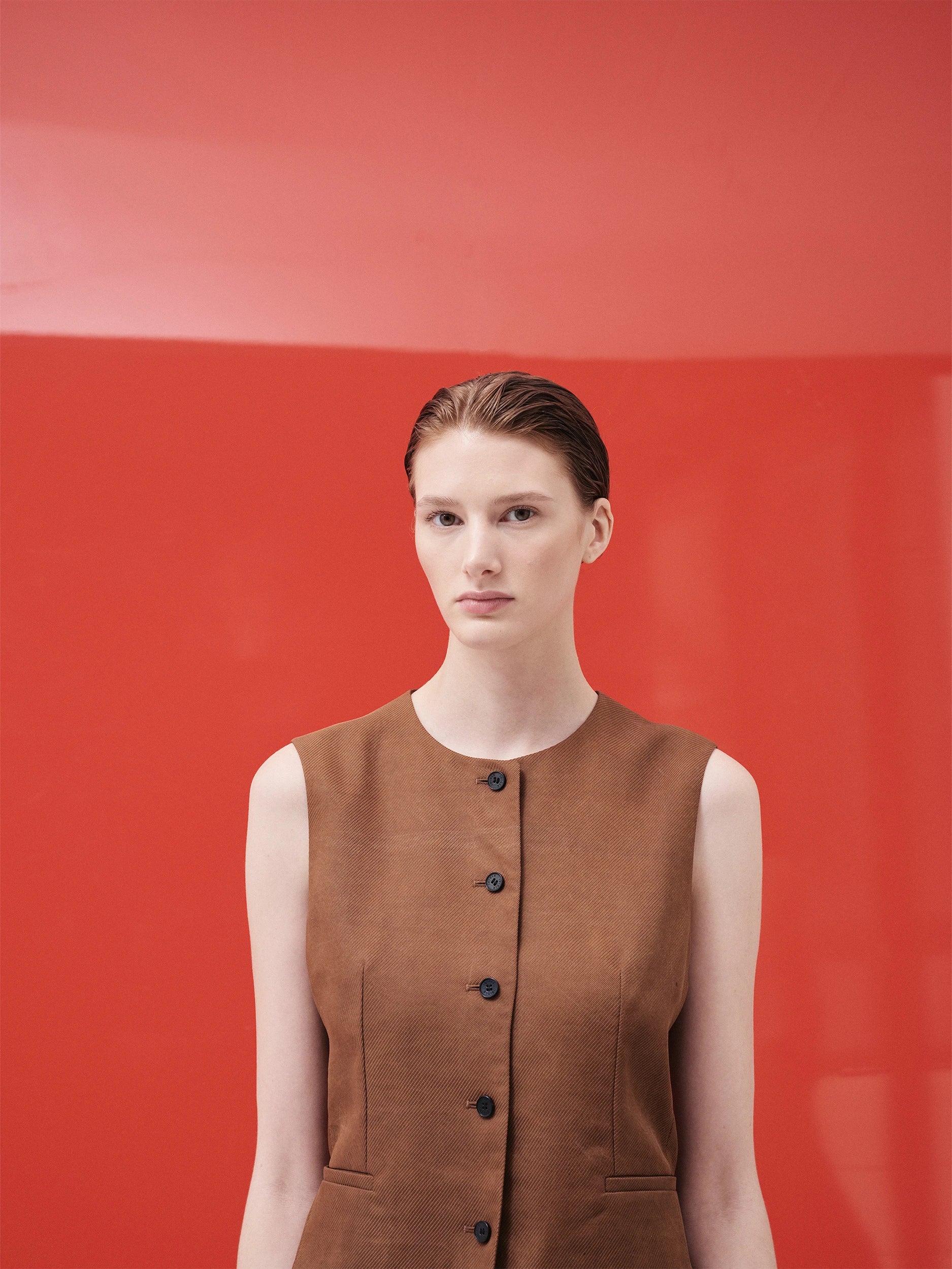 Model in the SS26 Canton Gauze collection wearing a camel-brown sleeveless collarless vest in structured woven gauze with a subtle textured weave, featuring dark horn buttons down the centre front and side pockets — a tailored, straight silhouette against a bold red background