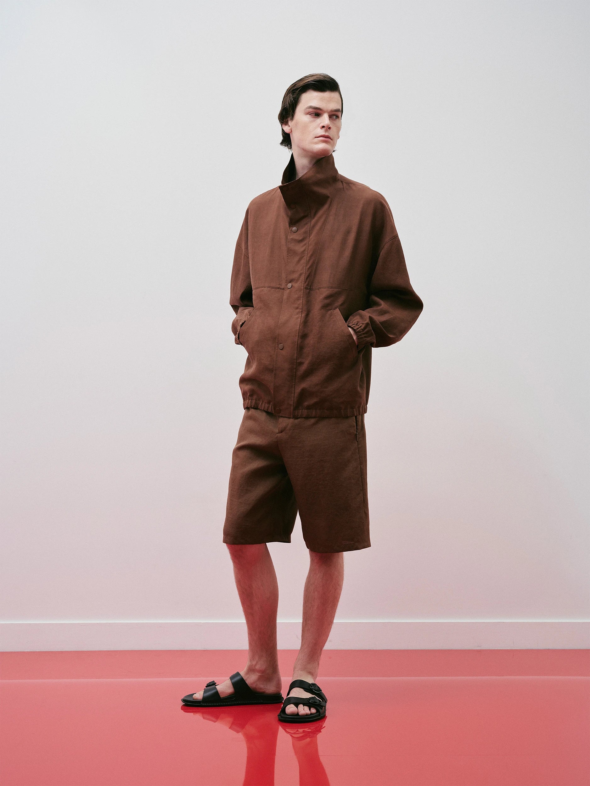 Male model in the SS26 Canton Gauze collection wearing a rich chocolate-brown blouson jacket in crisp, lightweight woven gauze with a stand collar, hidden placket, chest pocket and elasticated hem, paired with matching wide-cut bermuda shorts — a relaxed, utilitarian silhouette styled with black leather sandals on a red floor against a neutral background