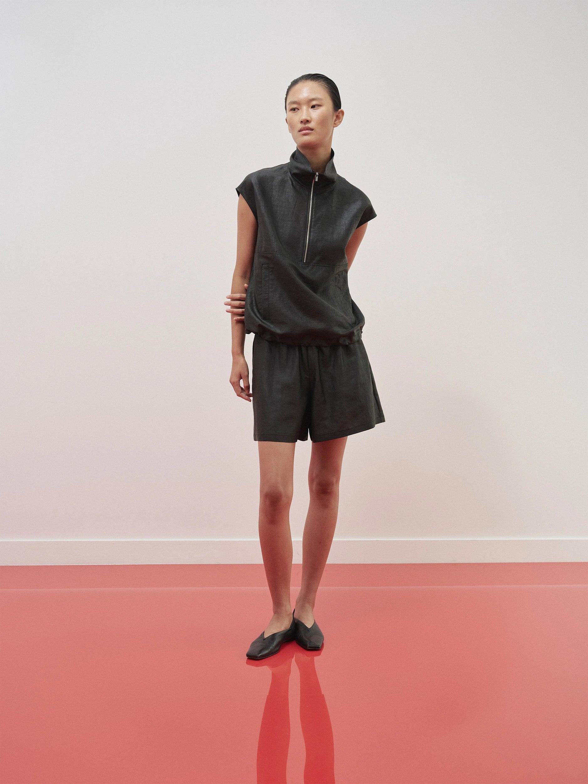 Model in the SS26 Canton Gauze collection wearing a dark black sleeveless zip-neck top in softly textured woven gauze with a relaxed drape, paired with matching charcoal wide-leg shorts with an elasticated waist — a casual yet refined monochromatic silhouette, styled with dark leather flat loafers on a red floor