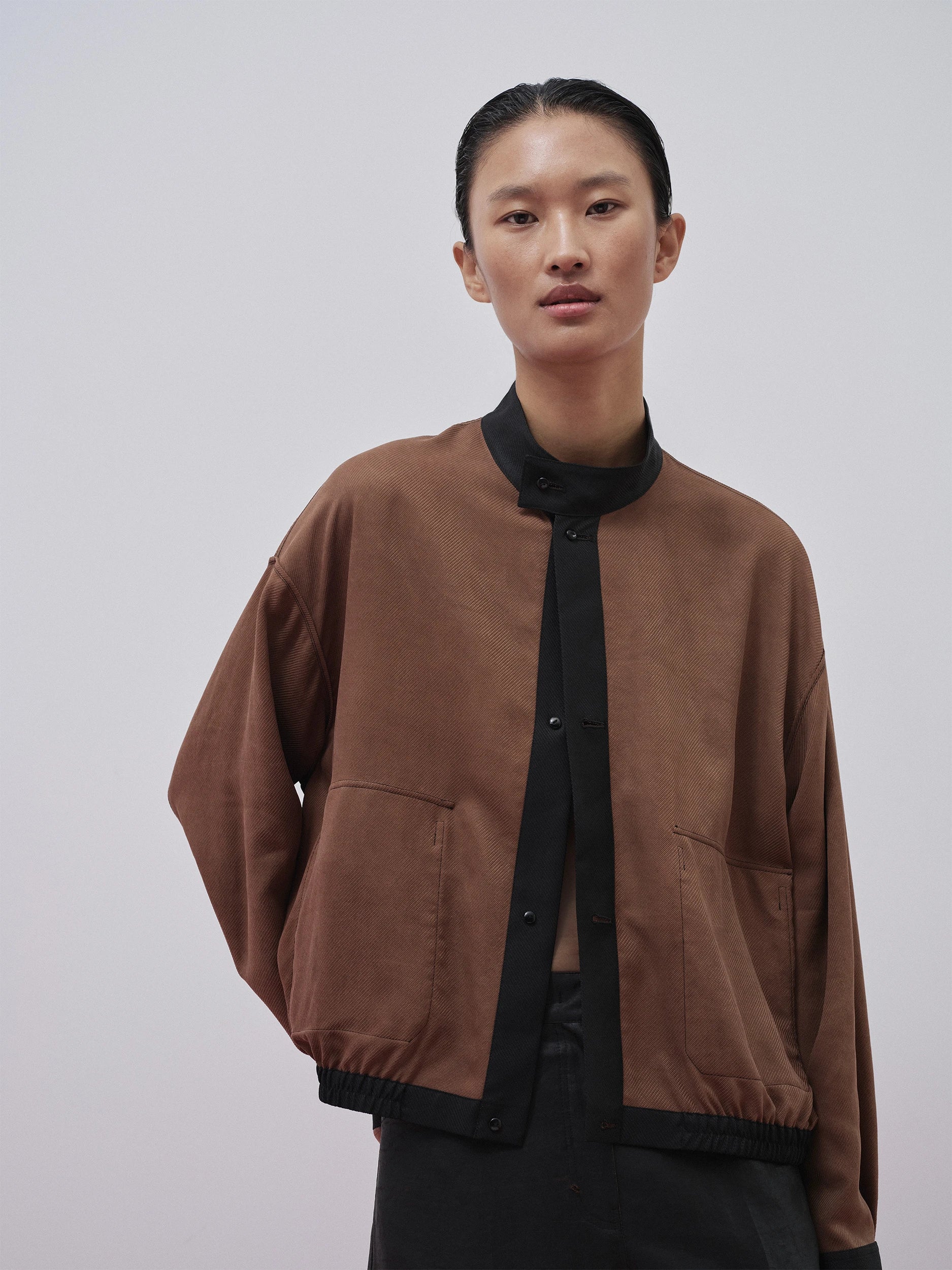 female model in the SS26 Canton Gauze collection wearing an oversized camel-brown bomber jacket in finely woven gauze with a subtle crosshatch texture, contrasting black ribbed collar, cuffs and hem, and two front patch pockets — layered open over a black mandarin-collar shirt and black skirt, creating a relaxed, cocoon-like silhouette against a neutral grey background