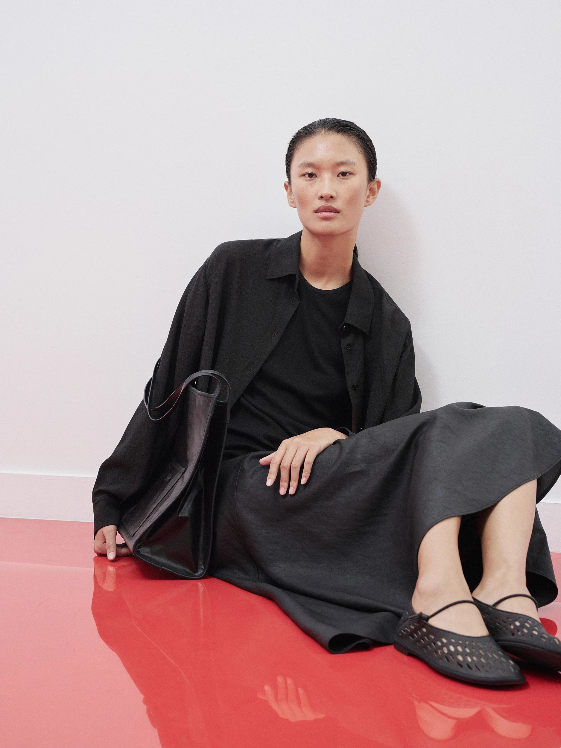Model in the SS26 Canton Gauze collection seated on a red floor, wearing an oversized black shirt in fluid lightweight gauze over a black crew-neck top and a wide black midi skirt with a soft drape — a voluminous, relaxed silhouette accessorised with a structured black leather tote and woven black flat shoes