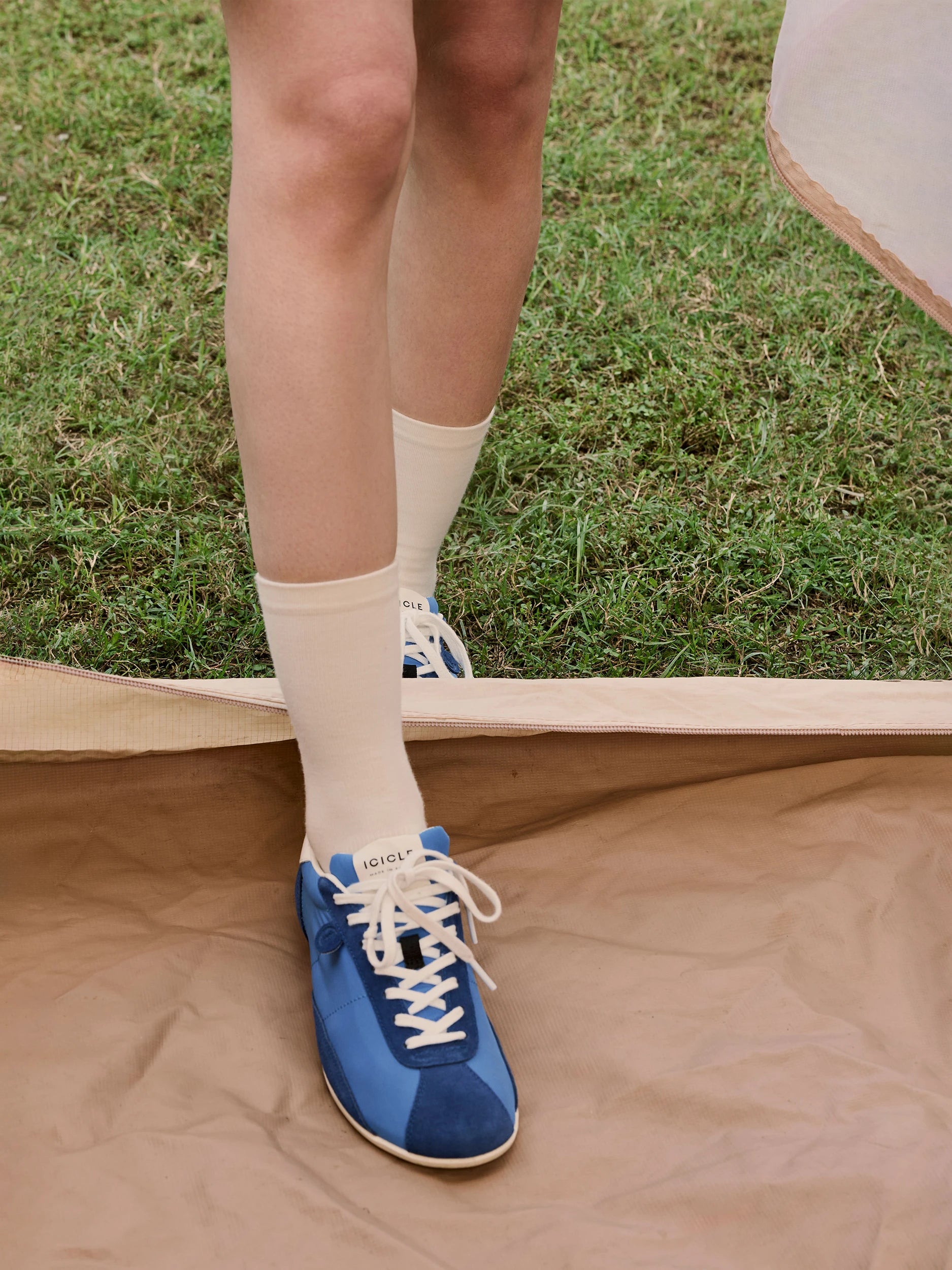 Close-up detail shot of a model's feet stepping between a grass lawn and a beige canvas groundsheet — wearing cobalt-blue retro lace-up sneakers in smooth leather and suede panels with a cream rubber sole and white lace-up fastening, branded with the Icicle logo on the tongue, styled with cream ribbed ankle socks with a small Icicle logo detail