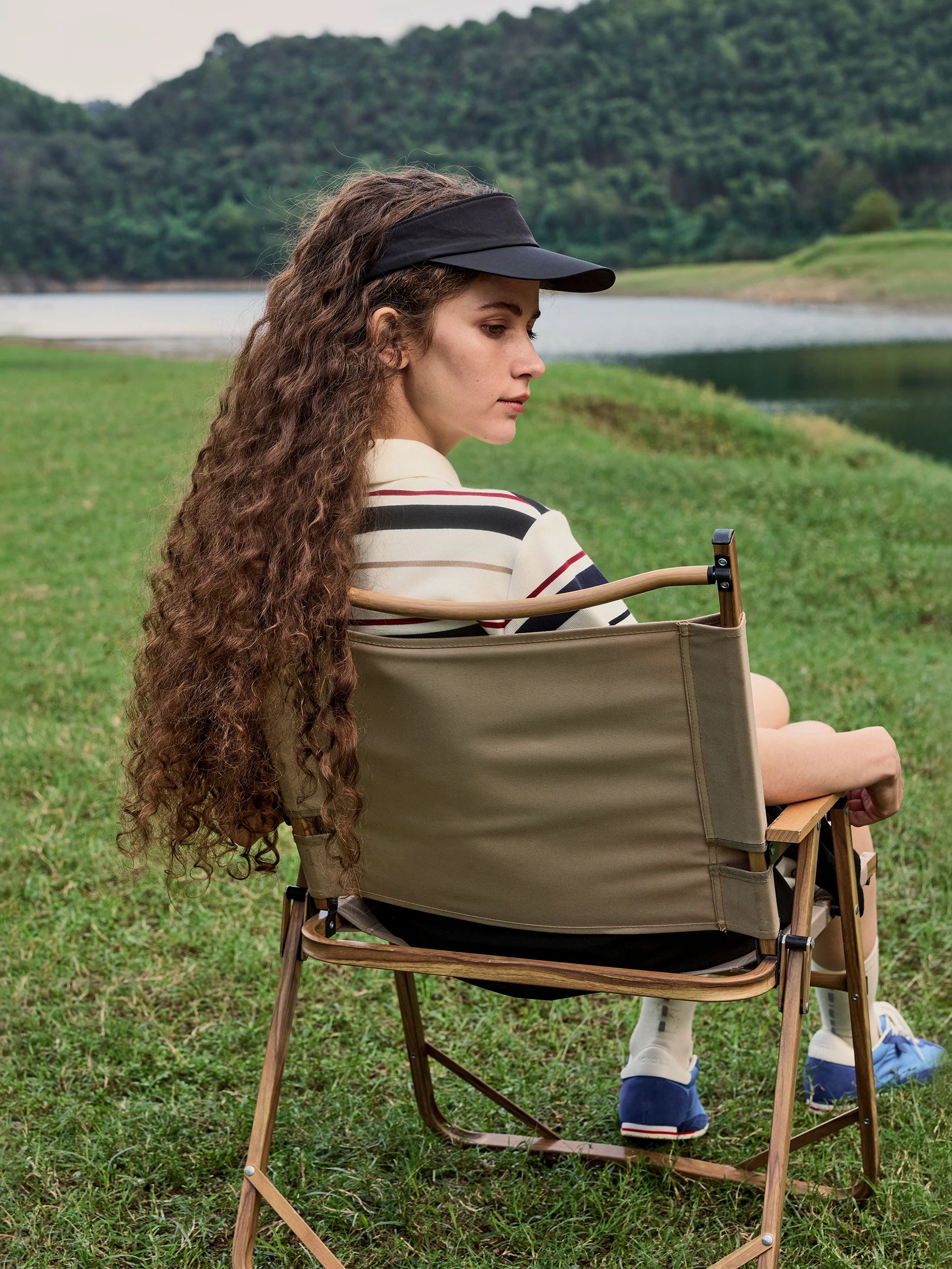 Female model seated in a folding wooden camp chair on a grassy lakeside, photographed in three-quarter profile — wearing a fitted short-sleeve polo shirt in smooth cotton jersey with cream, black and red horizontal stripes and a relaxed, slightly boxy silhouette, paired with black shorts, a black sun visor and blue-and-white retro canvas sneakers — a casual, sporty-chic outdoor look against a serene green hillside and lake backdrop