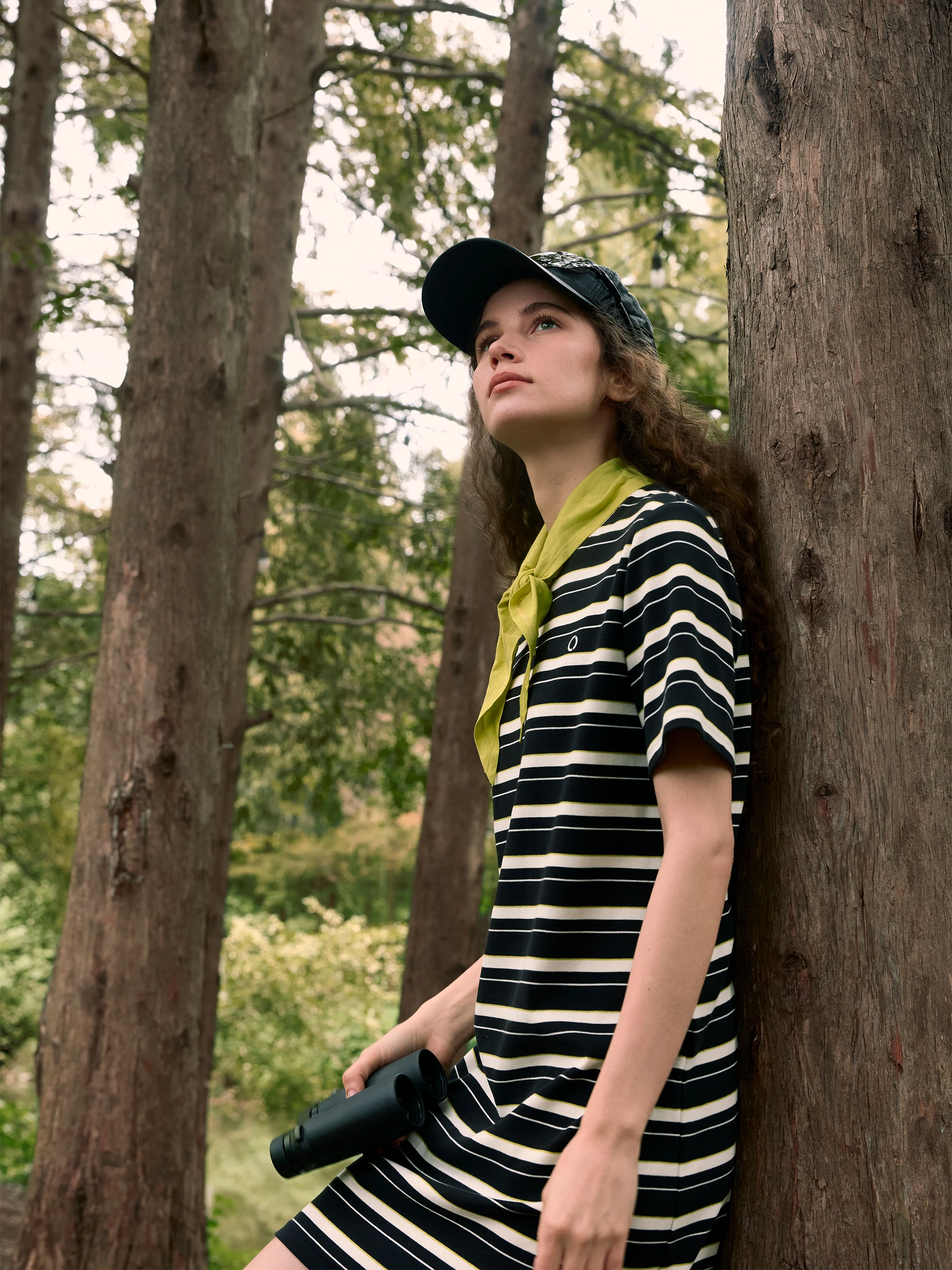 Female model leaning against a tall pine tree in a forest, gazing upward — wearing a short-sleeve midi dress in soft cotton jersey with bold black-and-cream horizontal stripes and a relaxed, straight silhouette, styled with a chartreuse-yellow neckerchief tied loosely at the collar, a dark navy printed bucket hat and holding a black monocular — a playful, nature-inspired look with a sporty-chic edge