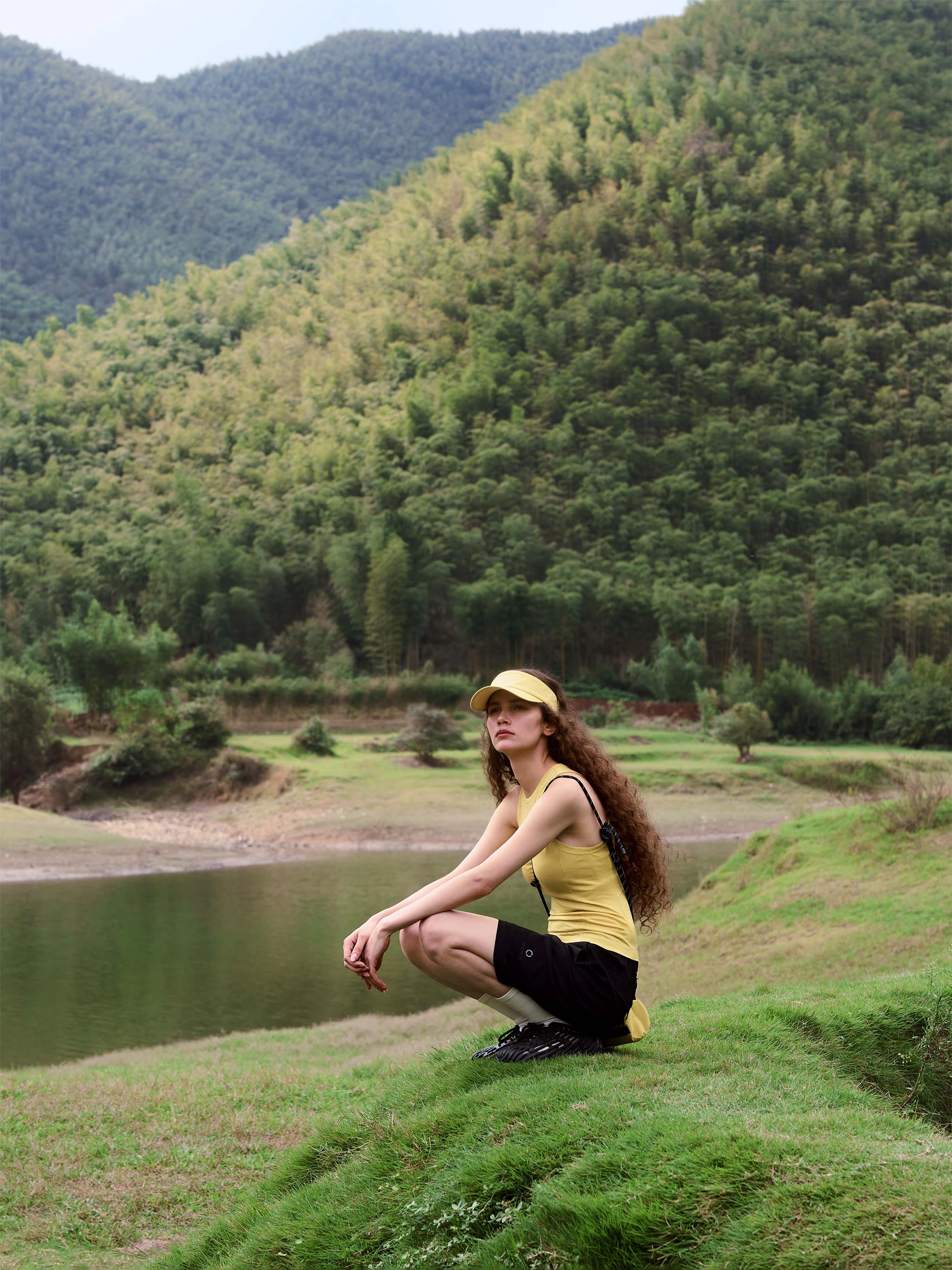 Female model crouching on a grassy hillside overlooking a tranquil mountain lake and lush bamboo-covered hills — wearing a fitted sleeveless tank top in smooth chartreuse-yellow jersey with a racerback silhouette, paired with tailored black bermuda shorts in structured fabric with a side cargo pocket, a matching yellow sun visor, a black crossbody bag and black chunky-soled hiking sandals — a sporty, functional outdoor look in a serene natural landscape