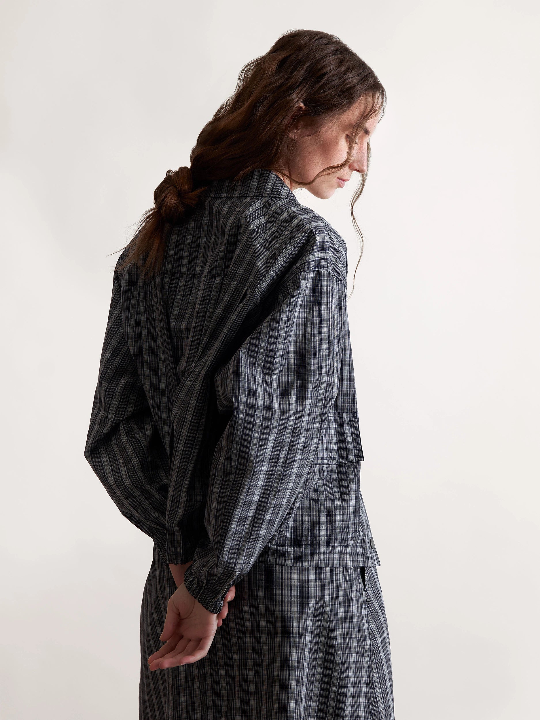 A model wears a checked cotton-silk jacket paired with a matching long skirt, highlighting the fluid textures and dark tonal variations of the silhouette.
