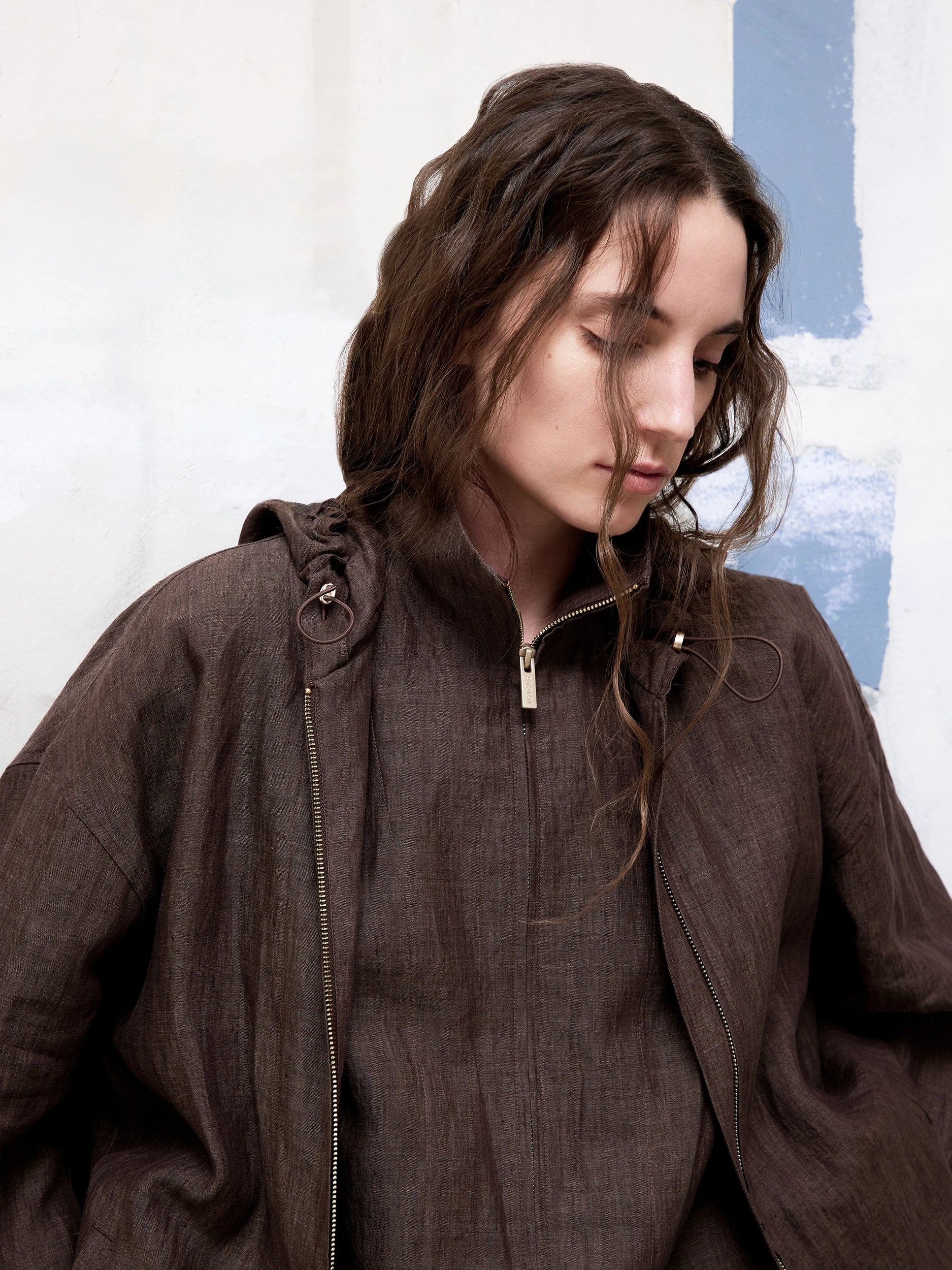 Close-up of a model wearing a textured dark brown silhouette with a hooded zip jacket against a light-toned and blue wall.