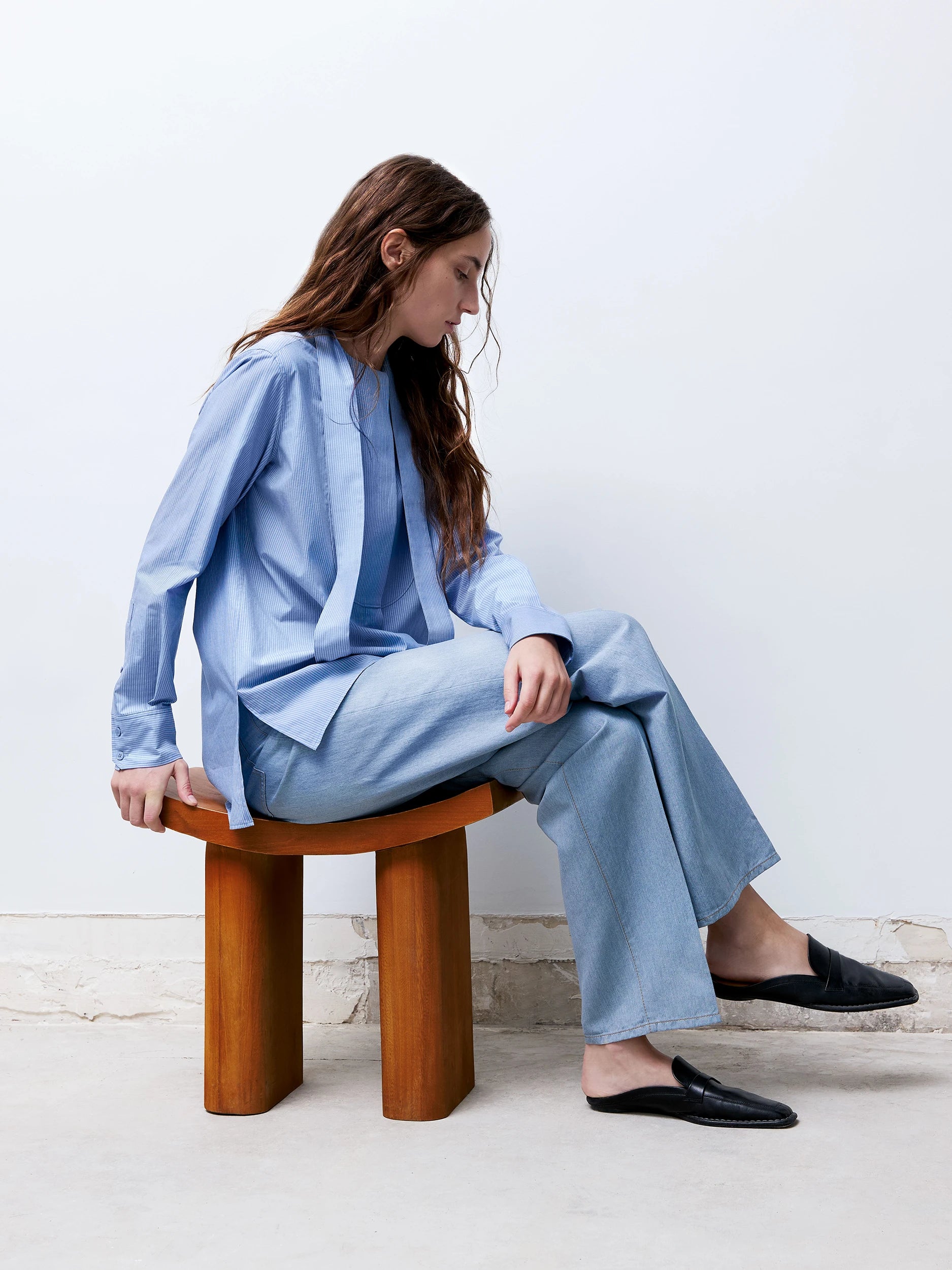 A model seated on a wooden stool wears a fluid light blue silhouette composed of a relaxed shirt and denim trousers in a minimalist setting.