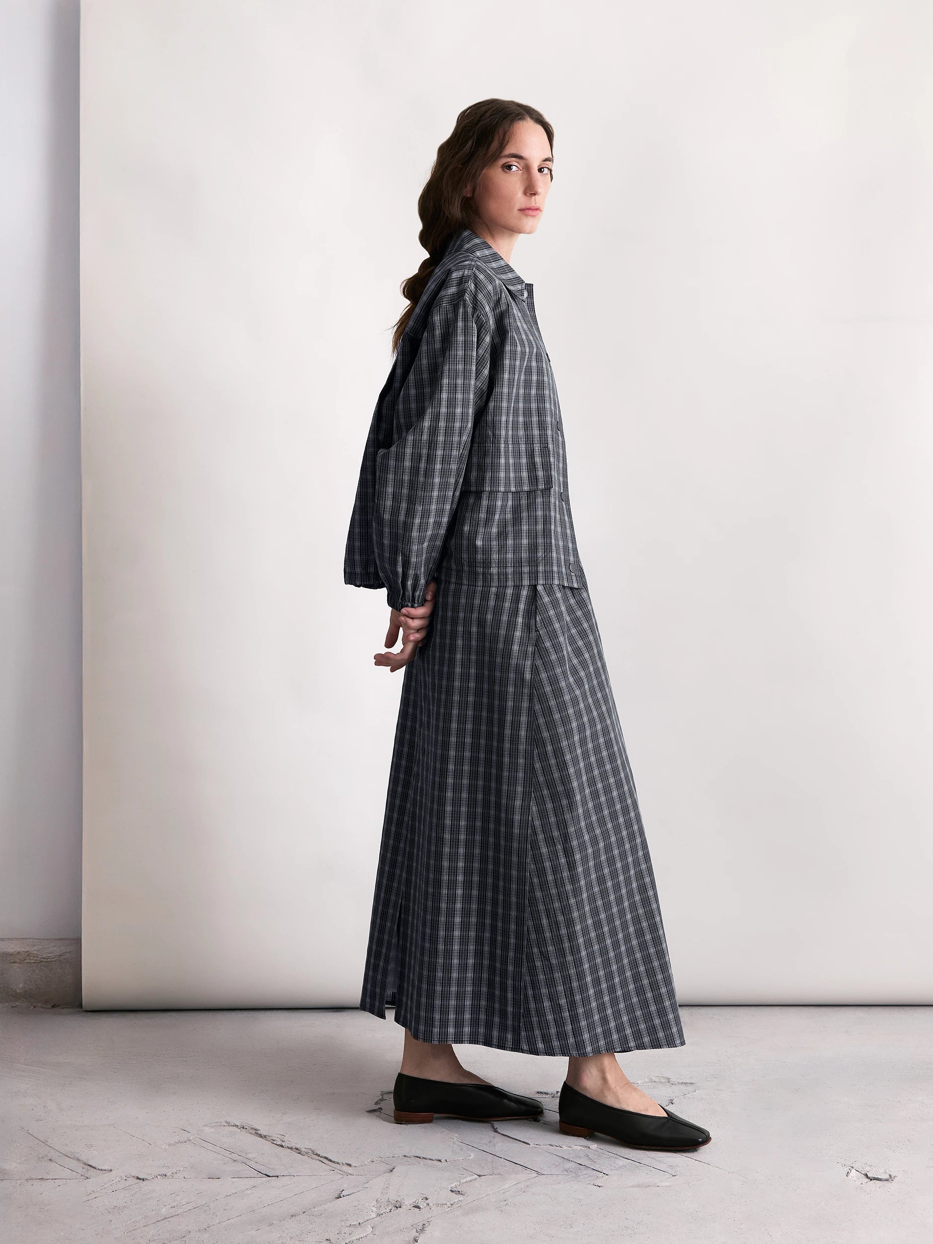 A model wears a checked cotton-silk jacket paired with a matching long skirt and black flats in a minimalist light-toned setting.