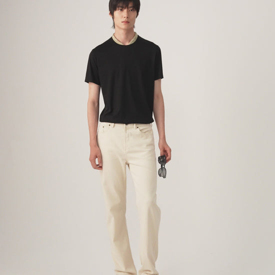Video of a male model stands against a plain light gray studio background wearing a black short-sleeve crew-neck t-shirt, off-white straight-leg trousers, and white sneakers, holding a pair of sunglasses in one hand.