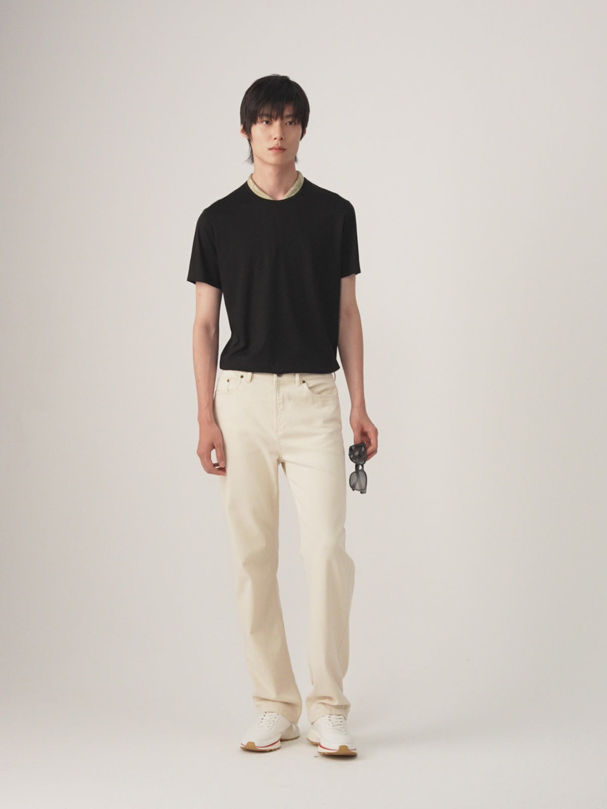 Video of a male model stands against a plain light gray studio background wearing a black short-sleeve crew-neck t-shirt, off-white straight-leg trousers, and white sneakers, holding a pair of sunglasses in one hand.