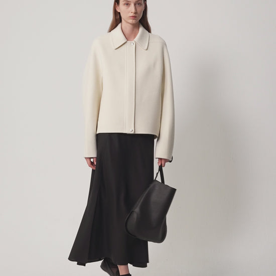 Video showing a woman wearing a cream-coloured double-sided wool and cashmere coat, paired with a black midi skirt and boots.
The model slowly turns to show the coat from different angles (front, side and back), revealing its long, flowing silhouette and natural, soft drape.
The movement highlights the coat's lightweight structure, the clean lines of the collar and the subtle texture of the undyed fabric.