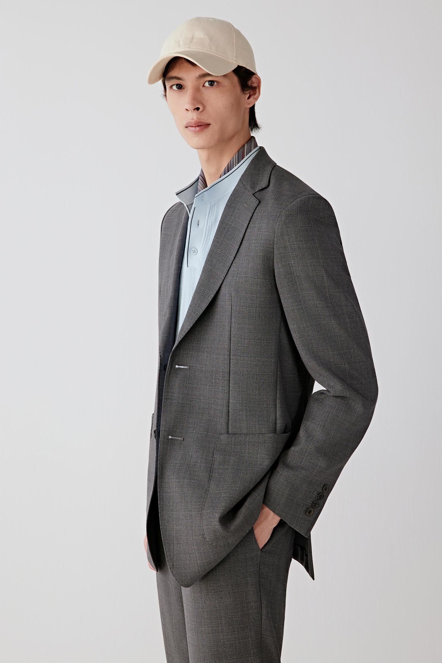 Video of a man standing against a plain light background, wearing a grey suit with a light blue shirt and white cap, holding a tennis racket and wearing white sneakers.