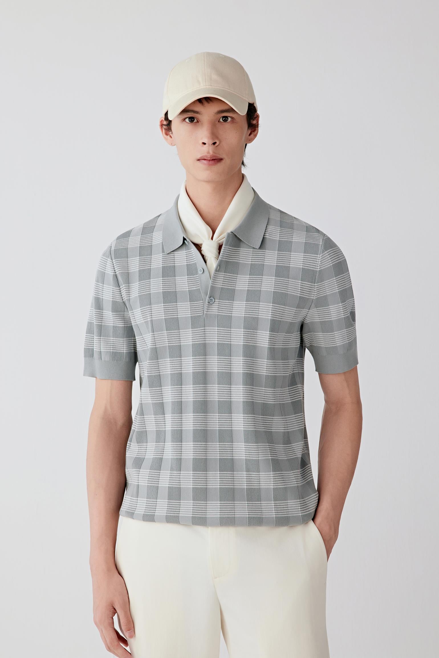Man wearing a light grey cotton knit polo-style pullover with a subtle checked pattern and relaxed straight silhouette, styled with off-white trousers and a cap. He turns and shifts between front, side and three-quarter views, revealing the lightweight, breathable cotton knit and its soft, fluid drape. The polo collar, short button placket and clean sleeve finishes remain consistently visible, while the fabric moves naturally with the body, showing ease through the shoulders and torso, and a balanced, non-r