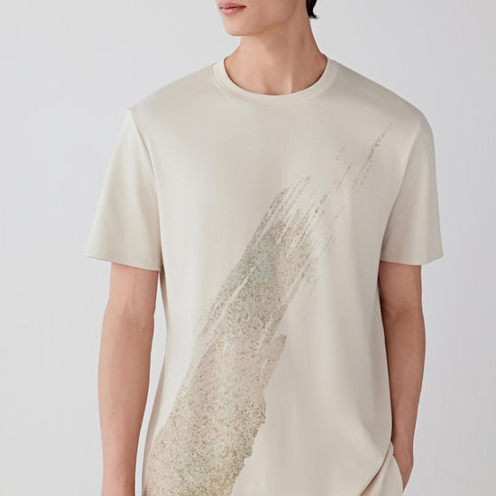 Man wearing a light nude cotton T-shirt with a relaxed straight cut and round neckline, styled with off-white trousers and a cap. He moves through front, side and angled views, showing the dense yet soft cotton jersey, breathable feel and natural drape. The clean neckline, short sleeves and integrated textured graphic on the front remain visible, while the fabric follows the body smoothly, emphasizing comfort, ease of movement and a balanced everyday silhouette.