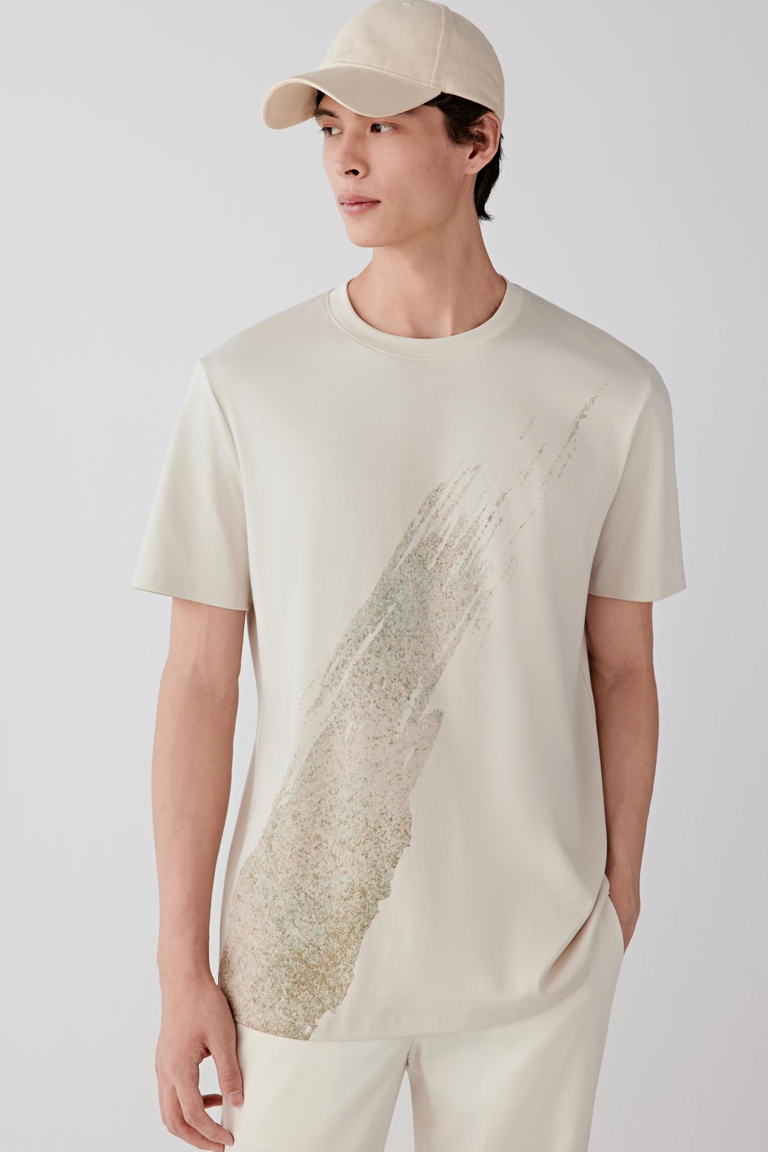 Man wearing a light nude cotton T-shirt with a relaxed straight cut and round neckline, styled with off-white trousers and a cap. He moves through front, side and angled views, showing the dense yet soft cotton jersey, breathable feel and natural drape. The clean neckline, short sleeves and integrated textured graphic on the front remain visible, while the fabric follows the body smoothly, emphasizing comfort, ease of movement and a balanced everyday silhouette.