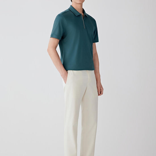 Man wearing a blue-green silk-cotton piqué polo shirt with a relaxed straight cut and zip collar, styled with off-white trousers and a cap. He moves between front, side and angled views, showing the breathable piqué knit, lightly structured feel and soft drape of the fabric. The short collar, zip fastening and clean sleeve finishes remain visible throughout, while the material follows the body naturally, highlighting ease through the shoulders and torso and overall comfort in motion.