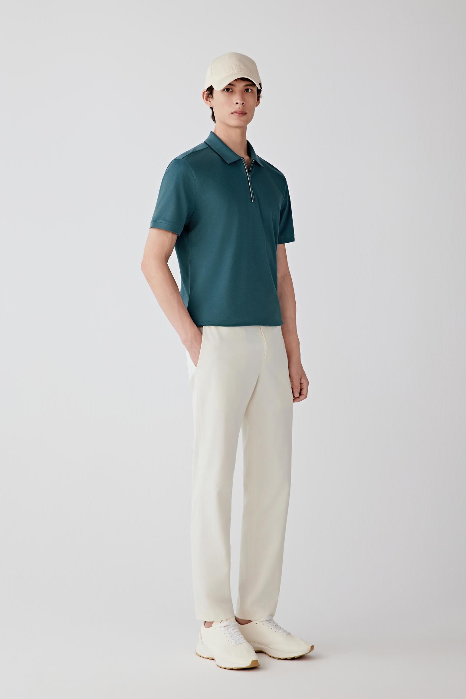 Man wearing a blue-green silk-cotton piqué polo shirt with a relaxed straight cut and zip collar, styled with off-white trousers and a cap. He moves between front, side and angled views, showing the breathable piqué knit, lightly structured feel and soft drape of the fabric. The short collar, zip fastening and clean sleeve finishes remain visible throughout, while the material follows the body naturally, highlighting ease through the shoulders and torso and overall comfort in motion.