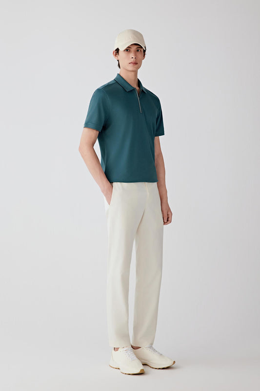 Man wearing a blue-green silk-cotton piqué polo shirt with a relaxed straight cut and zip collar, styled with off-white trousers and a cap. He moves between front, side and angled views, showing the breathable piqué knit, lightly structured feel and soft drape of the fabric. The short collar, zip fastening and clean sleeve finishes remain visible throughout, while the material follows the body naturally, highlighting ease through the shoulders and torso and overall comfort in motion.