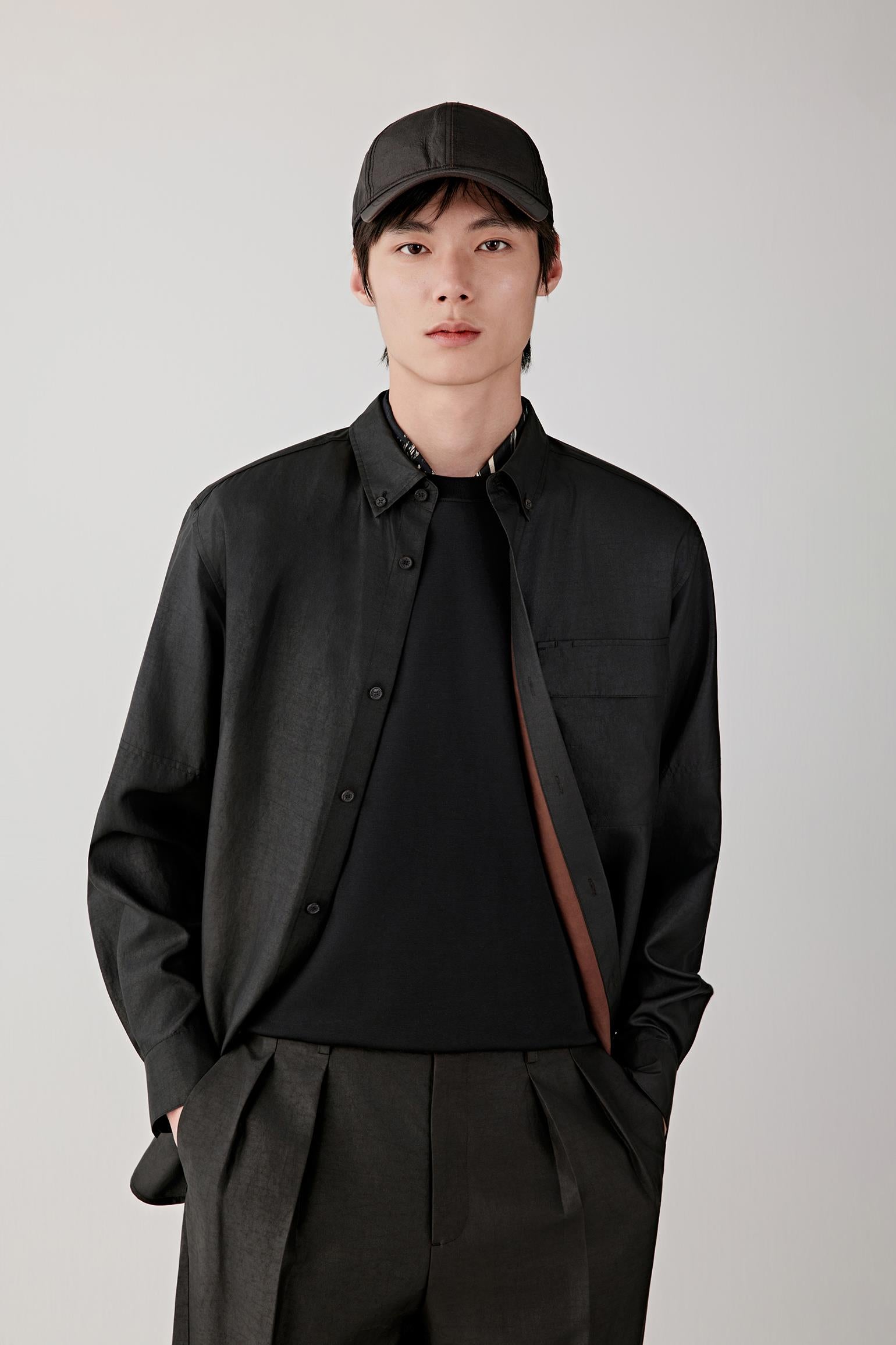 Man wearing a deep black Gambiered Canton gauze silk shirt with a relaxed straight cut and classic collar, styled with matching trousers. He moves through front, three-quarter and side views, showing the fluid drape, lightweight structure and naturally textured surface of the silk. The fabric shifts with light, revealing a subtle sheen, irregular grain and soft movement, while the clean lines, buttoned front and relaxed fit remain consistent throughout.