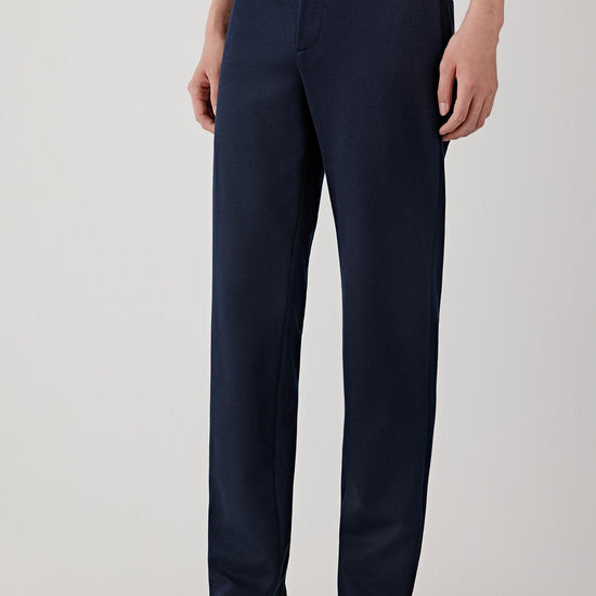 Man wearing navy ramie cotton and jacquard jersey trousers with a straight-leg cut and mid-rise waist, styled with a white T-shirt and cap. He moves through front, side and slight turning views, showing the clean vertical line, lightweight structure and soft drape of the fabric. The trousers fall straight from the hips with a natural, fluid movement, highlighting the subtle textured surface, comfortable fit and ease of motion.