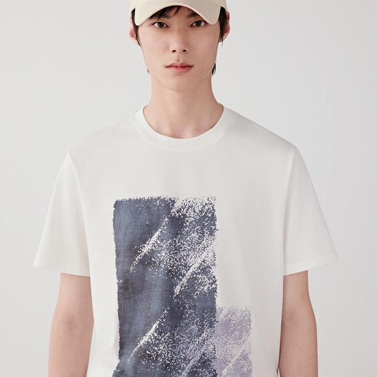 Man wearing a cream cotton jersey T-shirt with a relaxed straight cut, round neckline and abstract front graphic, styled with dark trousers and a cap. He moves between front, side and angled views, showing the lightweight, breathable jersey fabric and smooth drape. The graphic print remains centered and flat, while the clean neckline, short sleeves and soft structure emphasize comfort, fluid movement and an easy everyday fit.