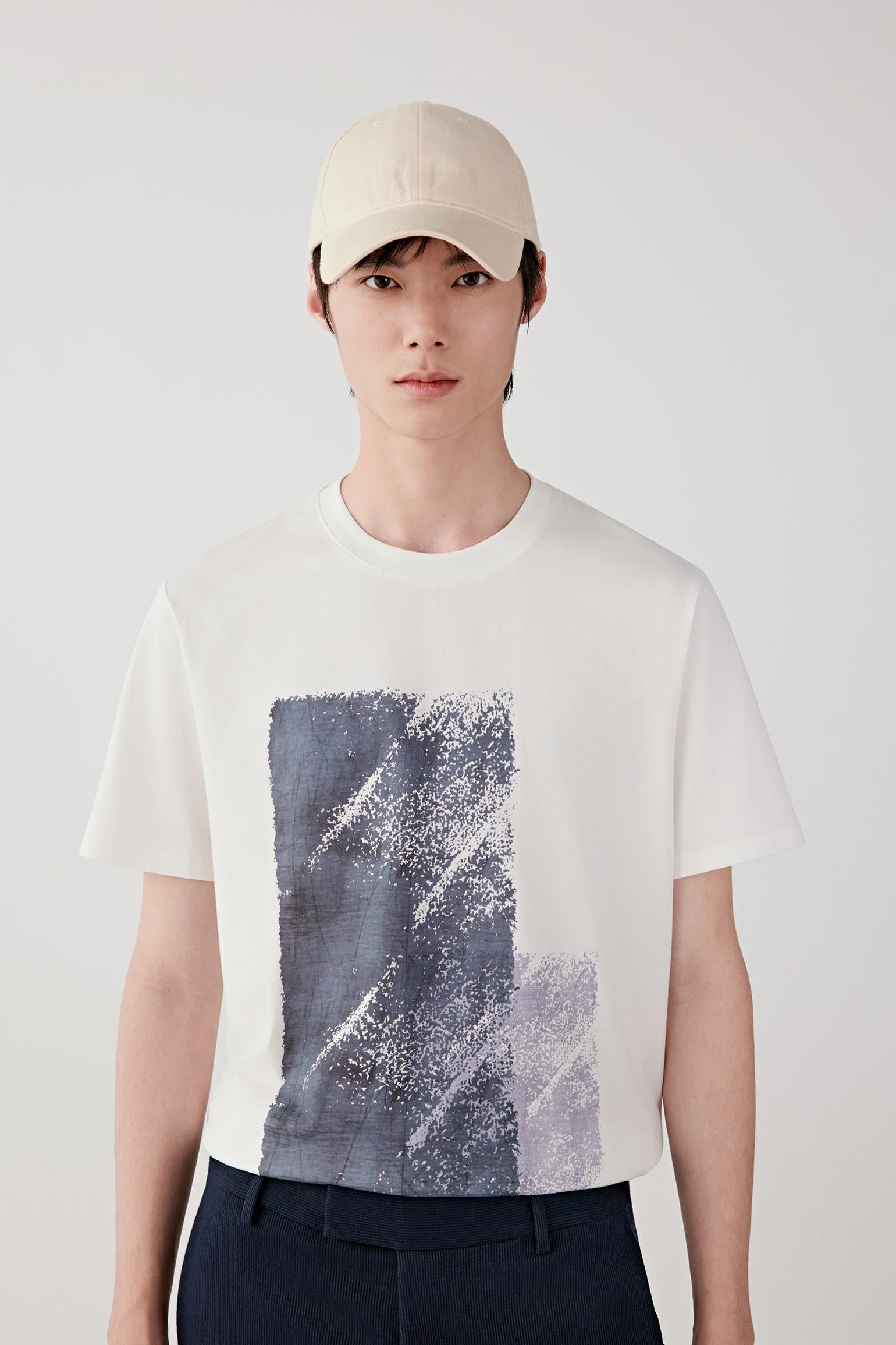 Man wearing a cream cotton jersey T-shirt with a relaxed straight cut, round neckline and abstract front graphic, styled with dark trousers and a cap. He moves between front, side and angled views, showing the lightweight, breathable jersey fabric and smooth drape. The graphic print remains centered and flat, while the clean neckline, short sleeves and soft structure emphasize comfort, fluid movement and an easy everyday fit.