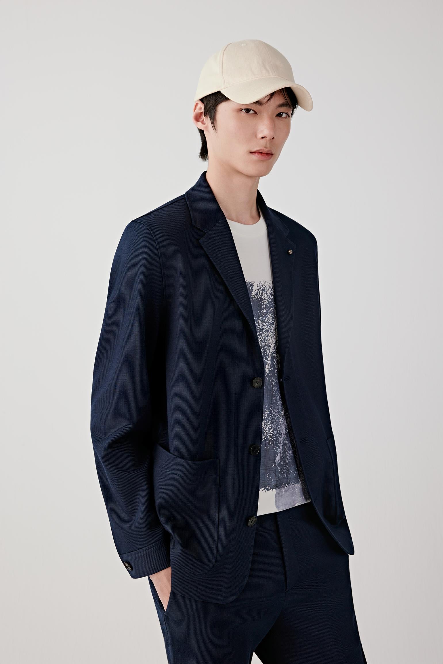 Man wearing a navy ramie cotton and jacquard jersey jacket with a straight, tailored cut and notched lapel, styled with matching trousers and a white T-shirt. He moves through front, three-quarter and side views, showing the lightweight structure, soft drape and subtle textured surface of the fabric. The jacket follows the body naturally, highlighting the clean lines, patch pockets and relaxed tailoring, with fluid movement emphasizing comfort and ease.
